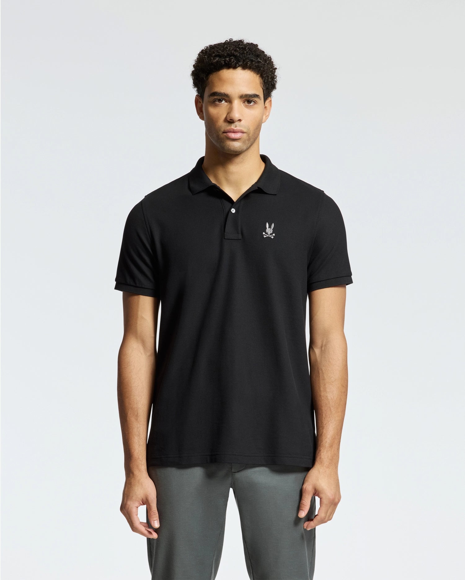 A man stands facing forward against a plain background, wearing a black Psycho Bunny MENS ALEXANDER PIQUE POLO - B6K101E200 with a logo on the left chest and gray pants.