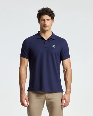 A man dressed in a Psycho Bunny Alexander Pique Polo (B6K101E200), featuring a small emblem on the left chest, stands against a plain light background. He has short curly hair and is wearing beige pants.