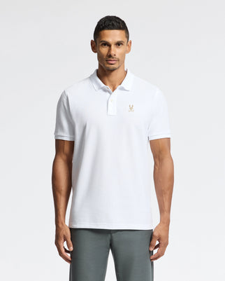 A man wearing a Psycho Bunny MENS ALEXANDER PIQUE POLO - B6K101E200, featuring four-way stretch fabric and a small embroidered logo on the left chest, stands against a plain white background. He has short dark hair and looks directly at the camera with his hands by his sides, radiating both style and durable breathability.