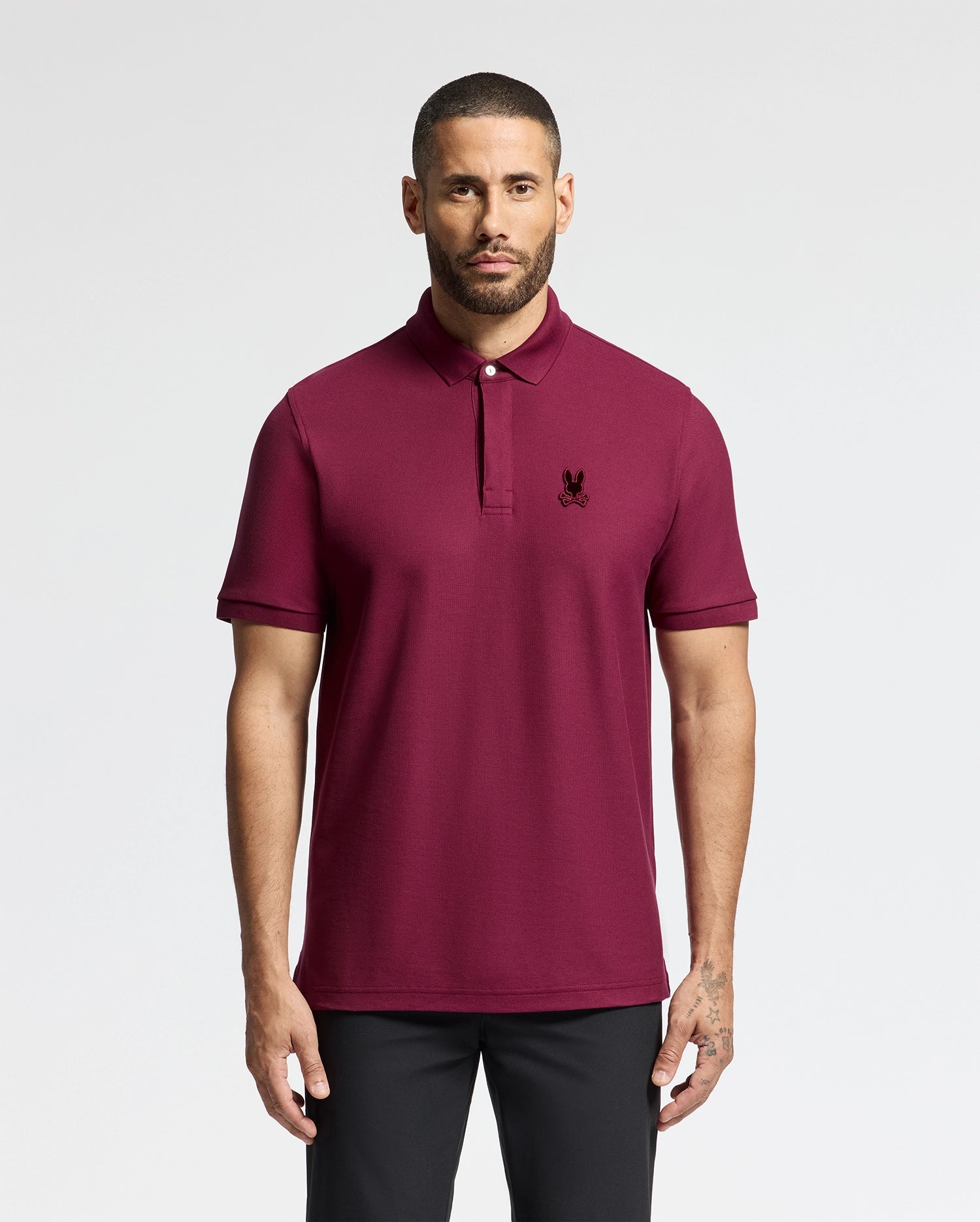 A man wearing a maroon Psycho Bunny MENS BRANSON PIQUE POLO (B6K108E200) with a small embroidered black logo on the chest is standing against a light gray background. He has a short haircut and a beard, with his hands relaxed at his sides, and he pairs it with black pants.