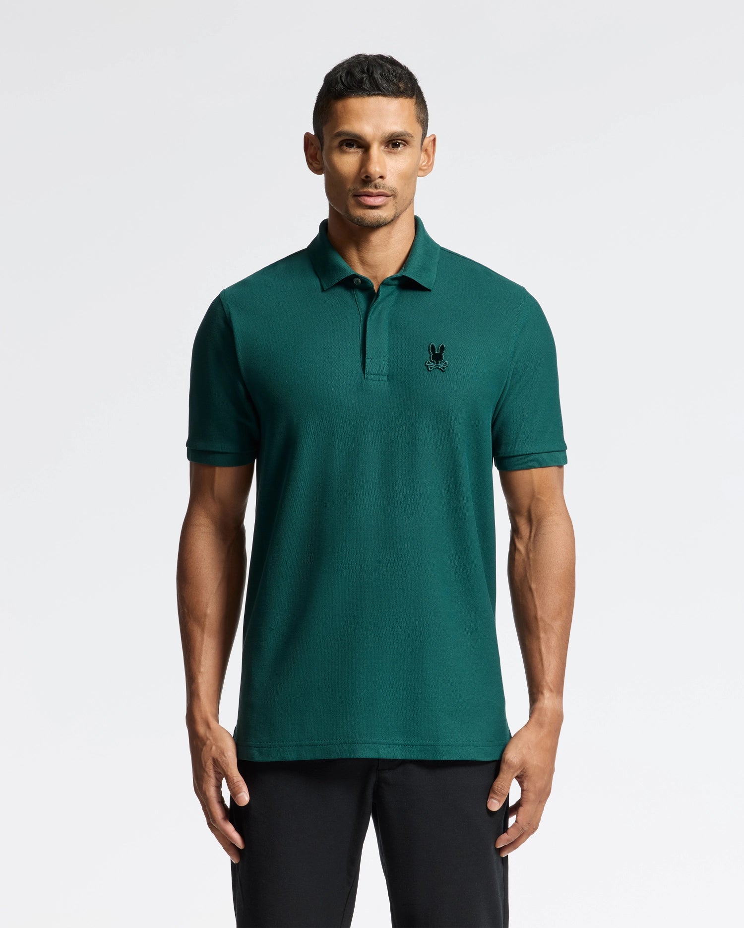 A man is wearing a dark green Psycho Bunny Men's Branson Pique Polo - B6K108E200, featuring a small logo on the left chest, standing against a plain white background. This polo shirt boasts a classic collar and short sleeves, making it ideal for any occasion.