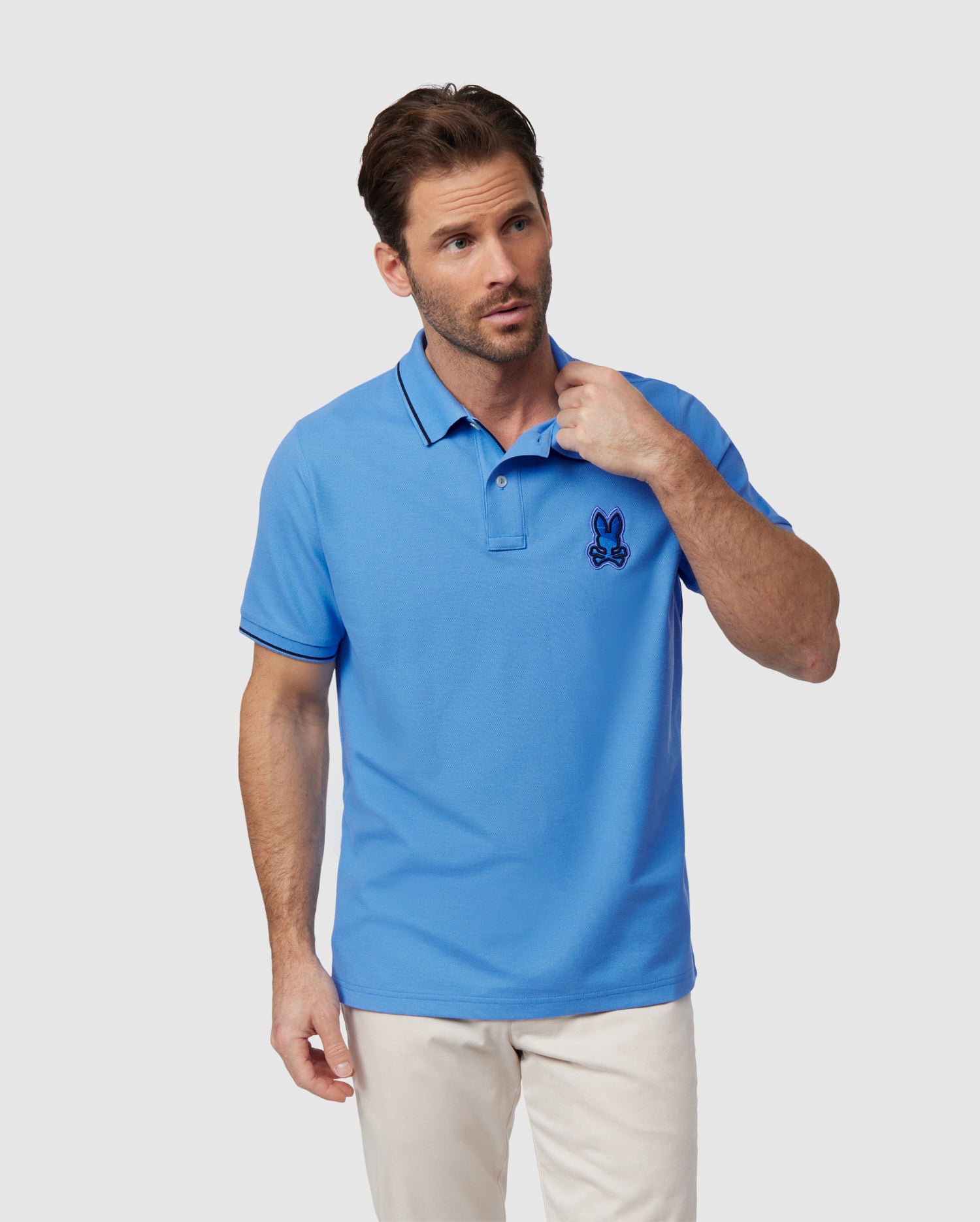 A man with short brown hair is standing against a plain background wearing a light blue Psycho Bunny MENS LENOX PIQUE POLO SHIRT - B6K138B200 made of soft Pima cotton. The shirt features an embroidered Bunny logo on the left chest. He is looking slightly to the side and has one hand near the collar of his shirt, paired with light-colored pants.