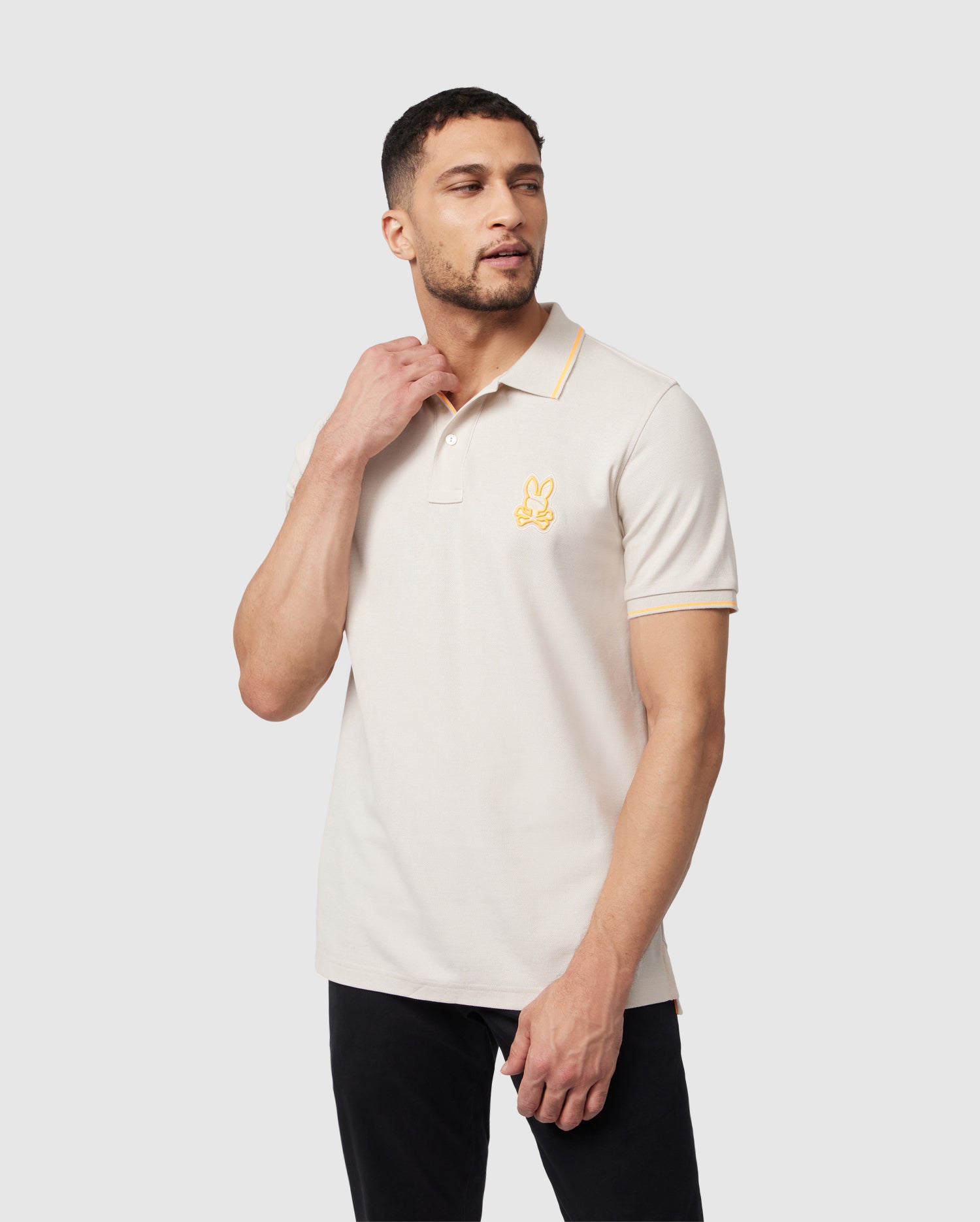 A man with short hair and a trimmed beard is posing in a light beige Pima cotton MENS LENOX PIQUE POLO SHIRT - B6K138B200 from Psycho Bunny with yellow contrasting tippings on the collar and sleeves. The shirt, complete with mother-of-pearl buttons, features an embroidered rabbit logo on the chest. He is touching his neck with his right hand and looking to his left.