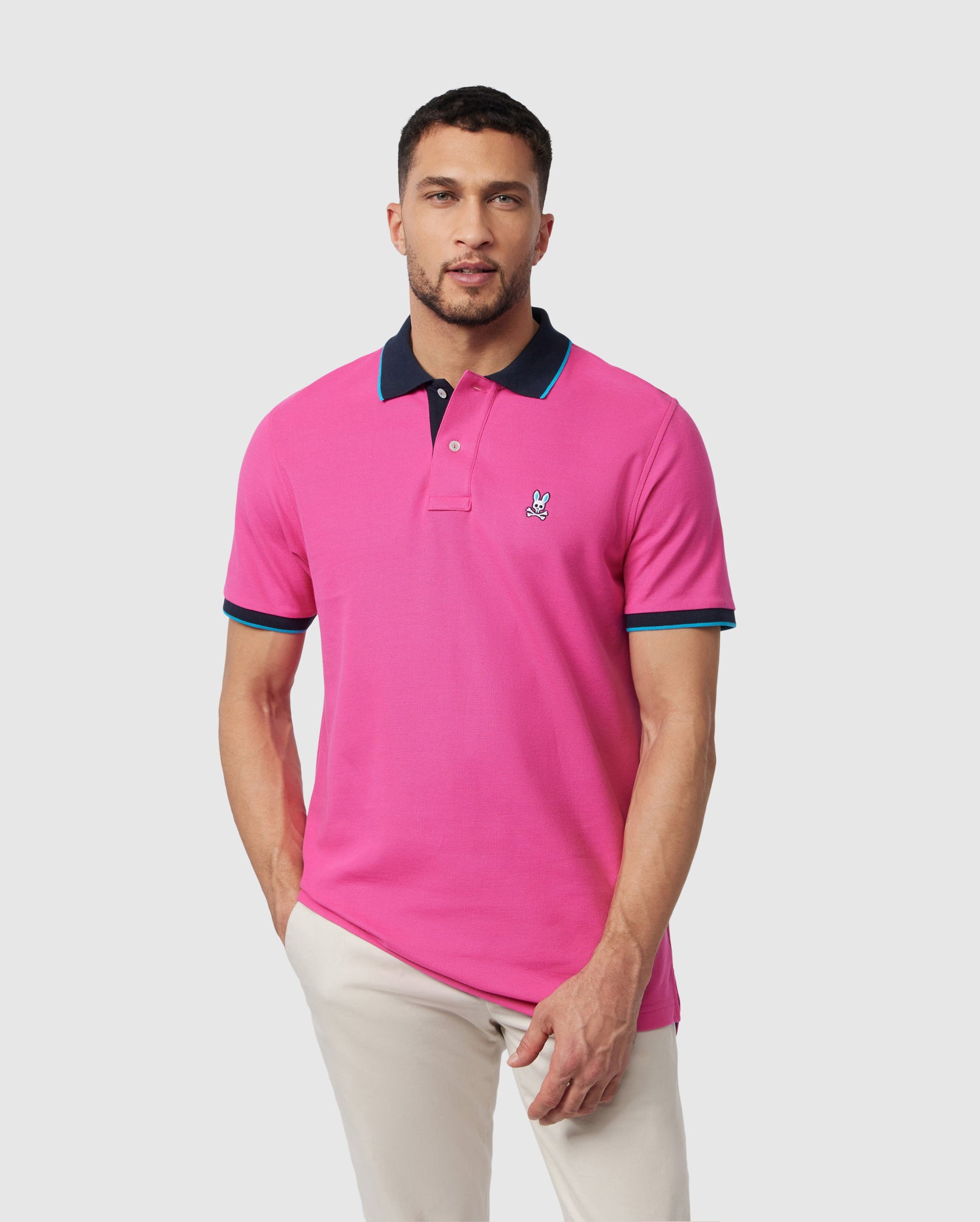 A man wearing a vibrant pink Psycho Bunny MENS TROY PIQUE POLO with blue accents on the collar and sleeves, posing against a plain background. He has short dark hair and a light stubble.