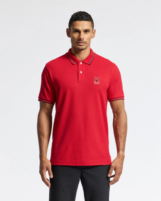 A person wearing a red Psycho Bunny MENS PIERCE PIQUE POLO - B6K201E200, featuring an embroidered bunny emblem on the left side of the chest, paired with black pants, stands against a plain white background.