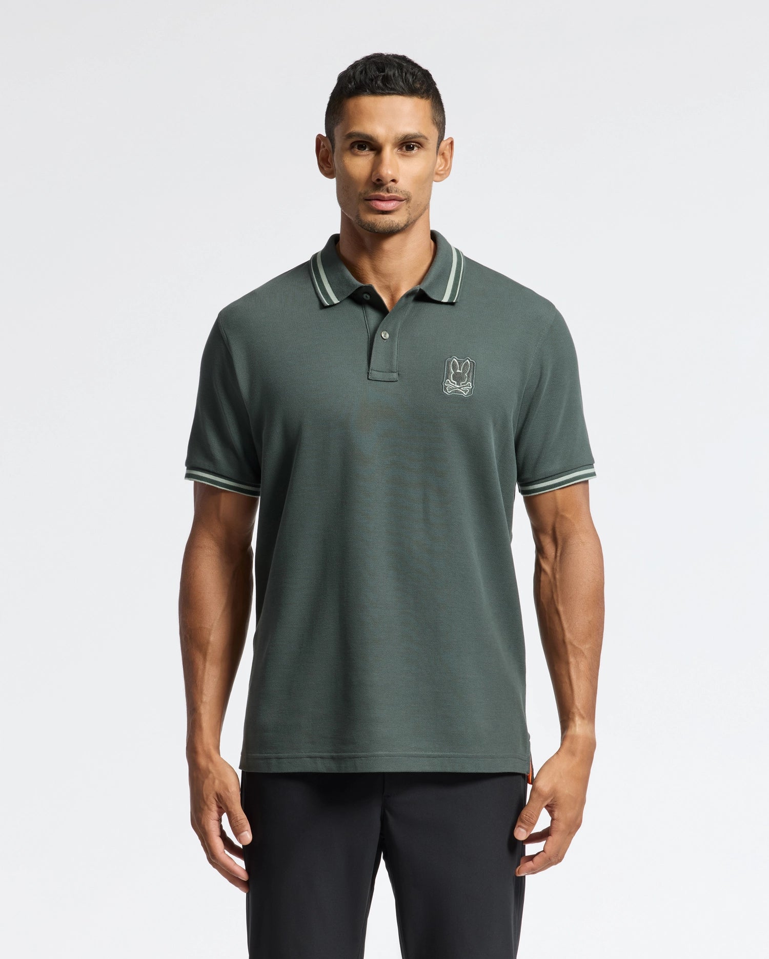 A man stands against a plain light background, wearing the Psycho Bunny MENS PIERCE PIQUE POLO - B6K201E200 in dark green, featuring white trim on the collar and sleeves. The polo is adorned with an embroidered Bunny logo on the chest and is stylishly paired with sleek black pants.