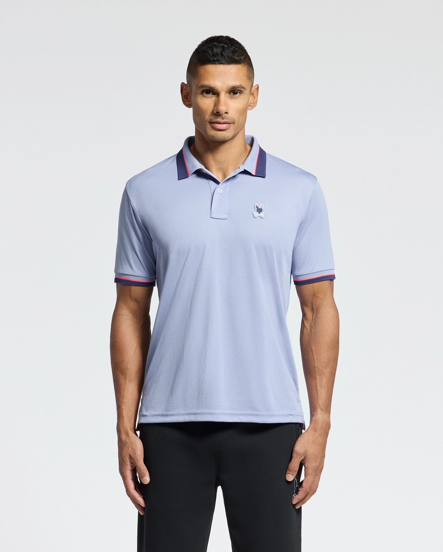 A man stands against a plain white background, wearing the Psycho Bunny MENS STRATTON SPORT PIQUE POLO - B6K202E200, a moisture-wicking light blue shirt with an embroidered logo, dark contrasting trim on the collar and cuffs, paired with black pants.