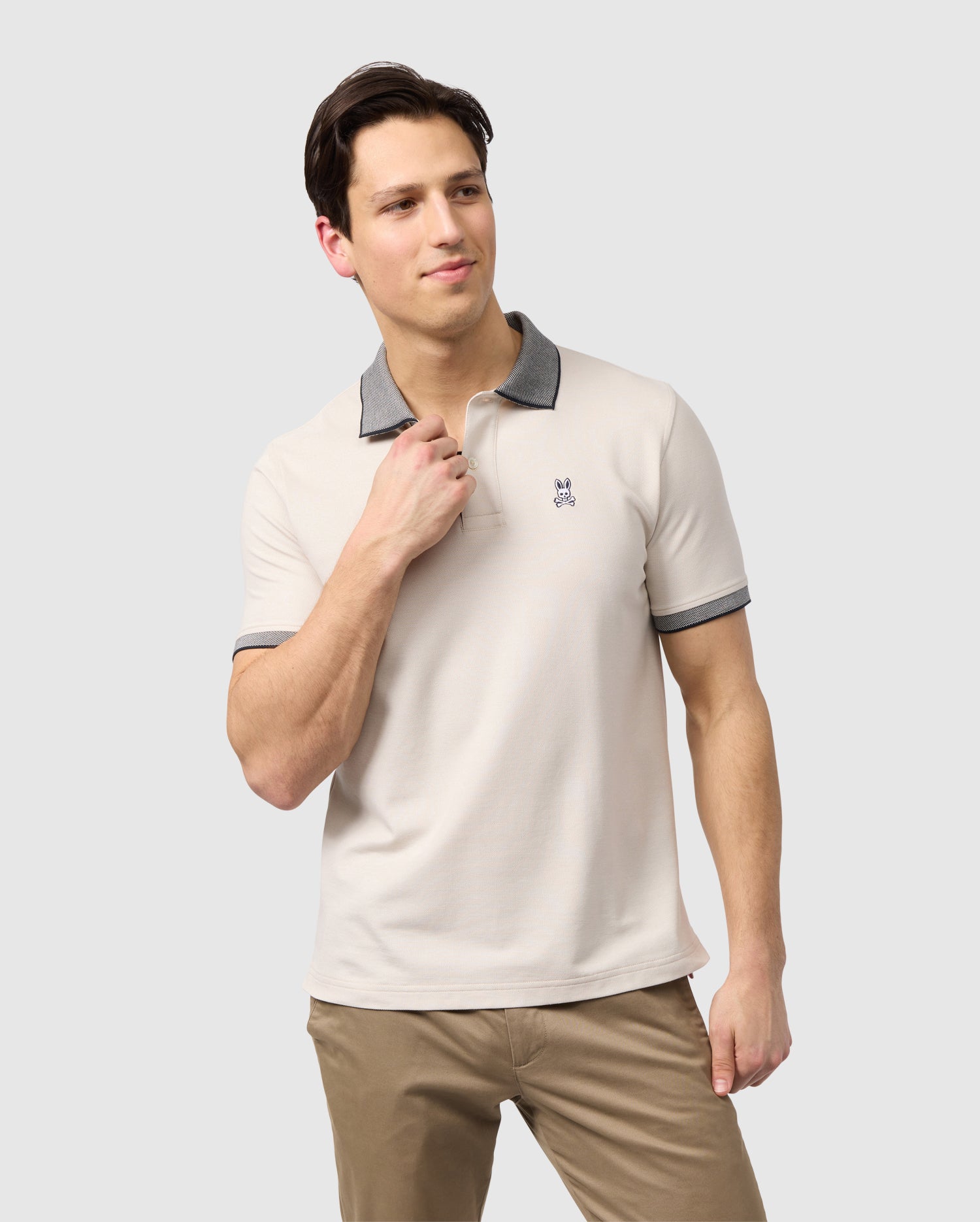 A man in a light beige Psycho Bunny Mens Southport Pique Polo Shirt and khaki pants, standing against a plain white background, adjusting his collar. He has short dark hair and is looking to his left.