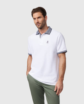 A man wearing a white Psycho Bunny Southport pique polo shirt (Product Code: B6K263B200) featuring a melange collar and sleeve edges, paired with light green pants. He stands with one hand in his pocket, looking off to the side against a plain background.