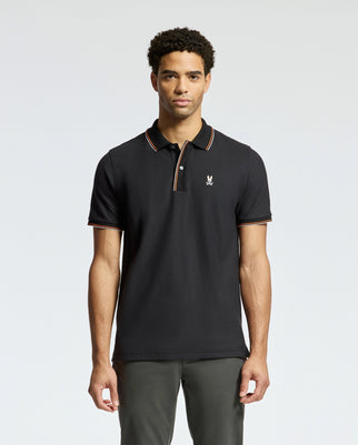 A person wearing the luxurious Psycho Bunny MENS BRANDON PIQUE POLO - B6K280E200, featuring orange and white trim and a small emblem on the left side, stands against a plain background. The individual with short curly hair pairs this premium quality polo shirt with dark pants.