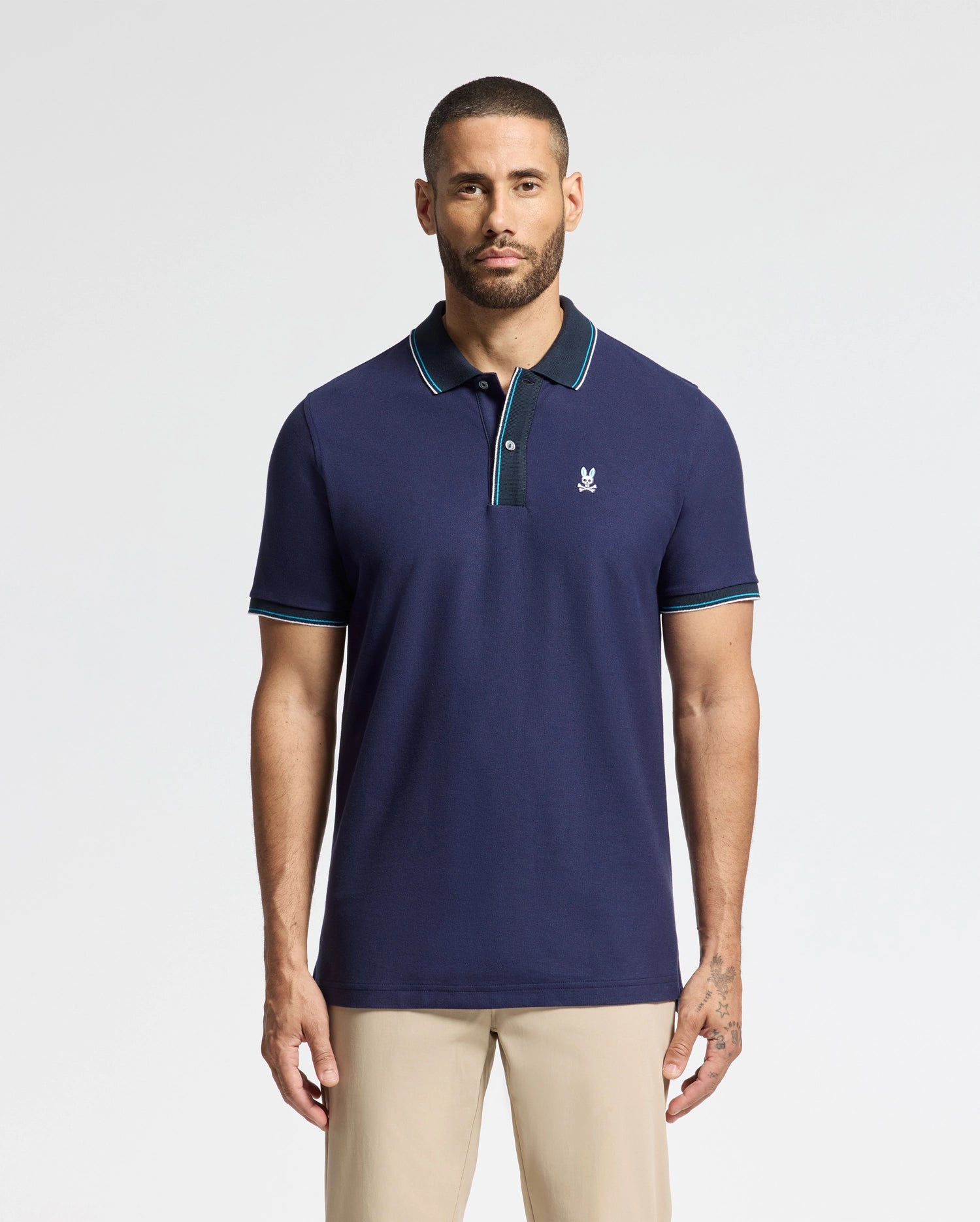 A man with short hair and a beard is wearing the Psycho Bunny MENS BRANDON PIQUE POLO - B6K280E200 in navy blue. The luxurious cotton polo features a small embroidered logo and has a green and white trimmed collar. He is standing against a plain white background, paired with beige pants.