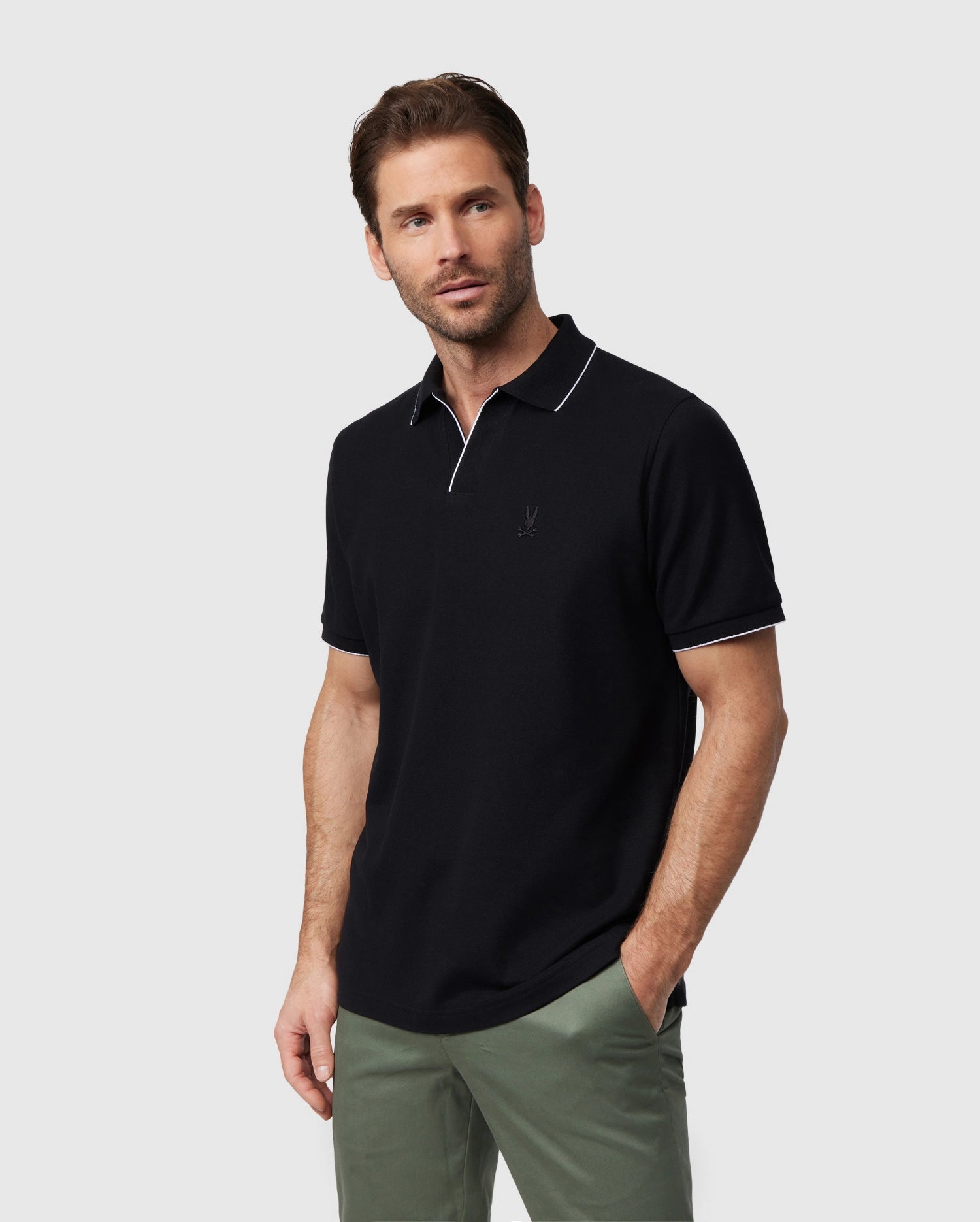 A man with short brown hair and a beard is wearing a black Psycho Bunny MENS EAST HILLS JOHNNY COLLAR POLO SHIRT - B6K331B200 with white trim on the Johnny collar and sleeves. He has one hand in the pocket of his light olive-green pants and looks to the side with a neutral expression. The background is plain gray.