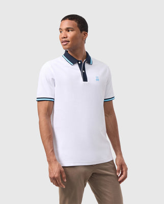 A man is wearing a standout Psycho Bunny MENS SALINA PIQUE POLO SHIRT - B6K379B200 made of Pima cotton, featuring a white base with blue stripes on the collar and sleeves. The shirt has a small blue logo on the chest. He is standing against a plain light gray background, also in light brown pants, looking to his right and smiling slightly.