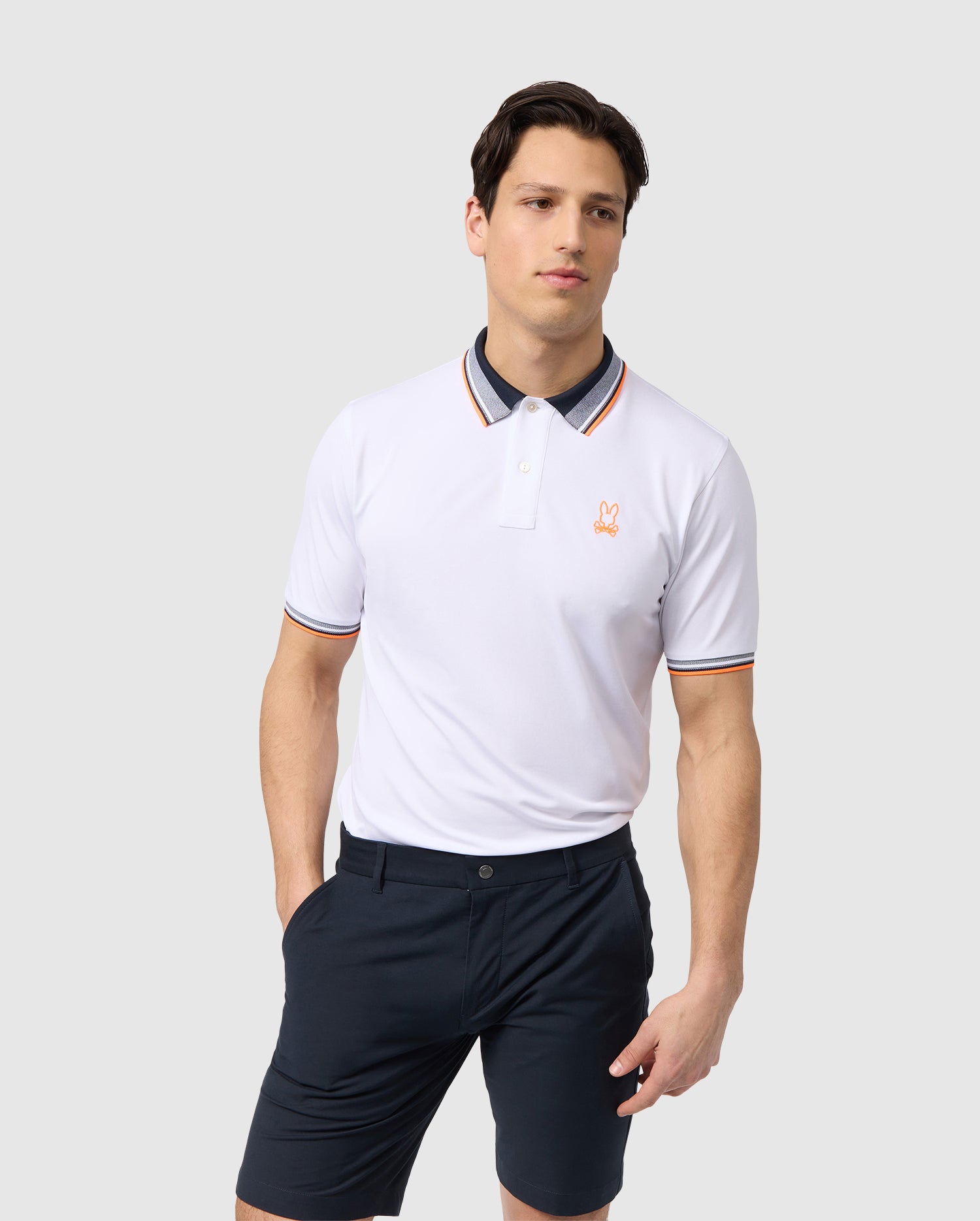 A man with short, dark hair is wearing a white Psycho Bunny MENS PORTLAND SPORT POLO - B6K397B200 featuring orange and black striped accents, an embroidered logo, and breathable mesh for added comfort. He pairs it with dark navy blue shorts. He is standing against a plain white background with one hand in his pocket and a neutral expression.