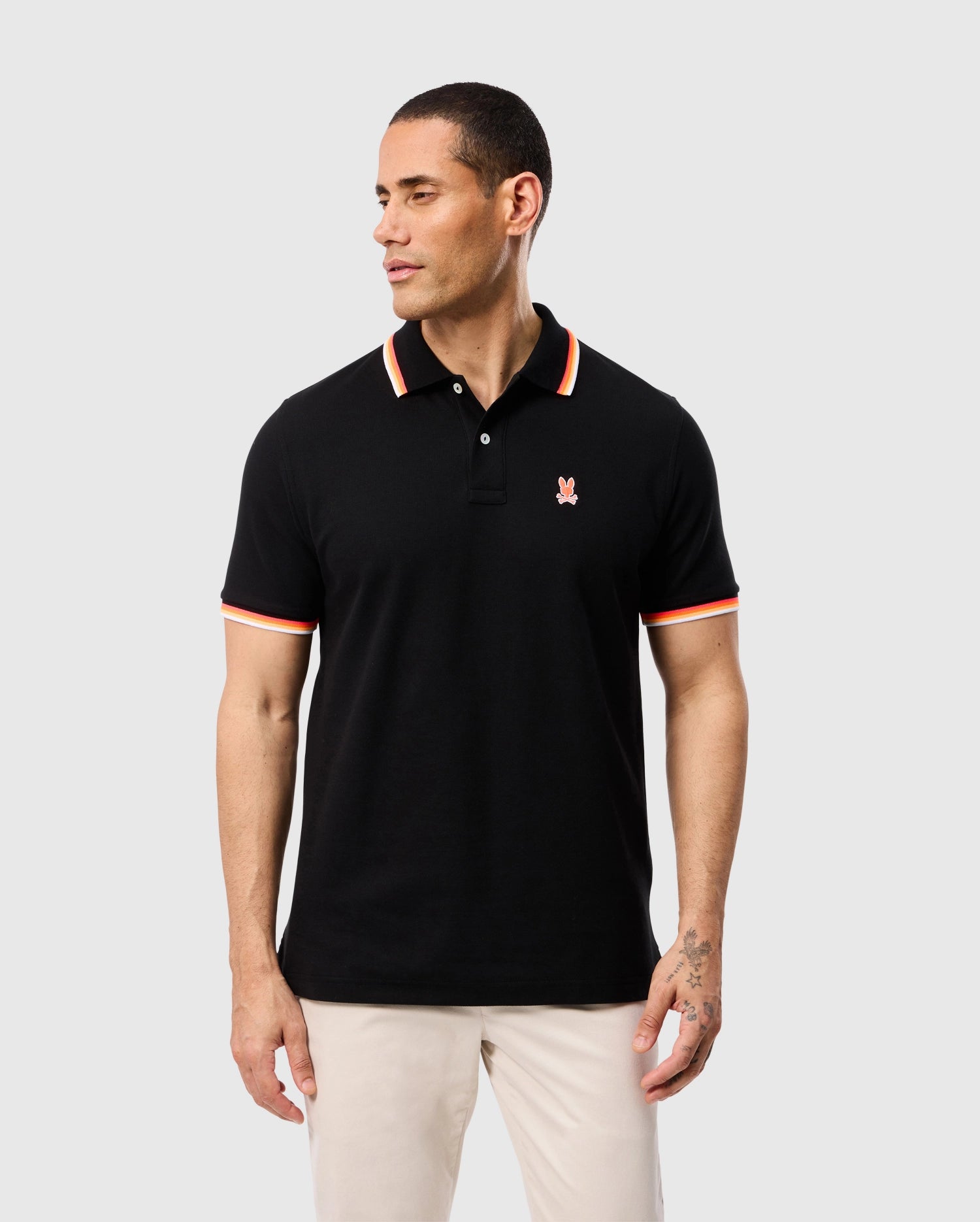 A man wearing a black Psycho Bunny MENS GRANBURY PIQUE POLO - B6K400C200 with red and white trim on the rib knit collar and cuffs stands against a plain gray background. The shirt, made from diamond-knit Pima cotton piqué, features a red embroidered logo on the left chest. He has short hair and is looking to his left while keeping his hands at his sides.
