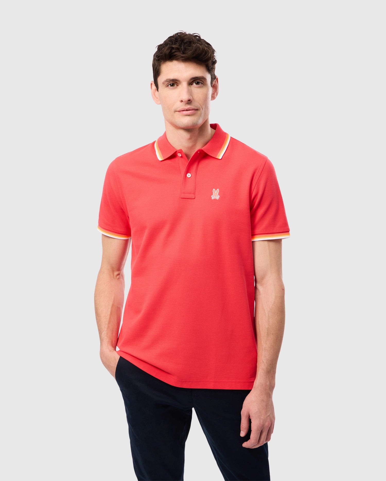 A person is wearing a Psycho Bunny MENS GRANBURY PIQUE POLO - B6K400C200 in bright red Pima cotton, featuring yellow and white trim on the collar and sleeves. The shirt has a small logo on the left chest. They have short dark hair and are standing against a plain, light gray background, also dressed in dark pants.