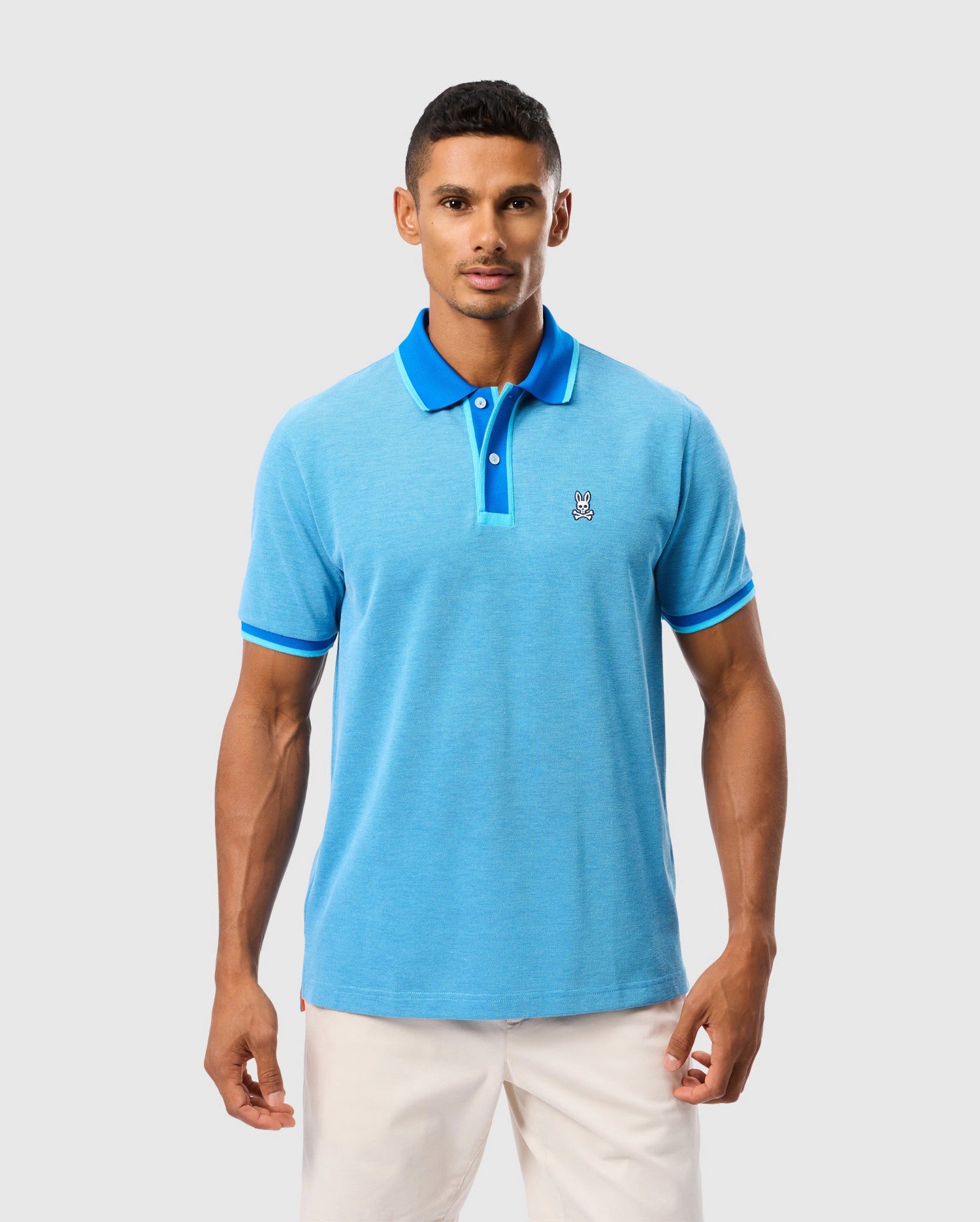 A man is wearing a light blue Psycho Bunny MENS WOODWAY OXFORD POLO - B6K405C200 with dark blue accents on the collar and sleeves. The Pima cotton-blend shirt features a two-button placket and has a small embroidered patch of a Bunny cartoon character on the left chest. He is also wearing white pants. The background is plain and gray.