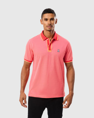 A man is wearing the Psycho Bunny MENS WOODWAY OXFORD POLO - B6K405C200, a bright pink short-sleeved shirt with orange trim on the collar and a classic two-button placket. The Pima cotton-blend polo features a small embroidered bunny logo on the left chest. He pairs it with black pants and stands against a plain light gray background.