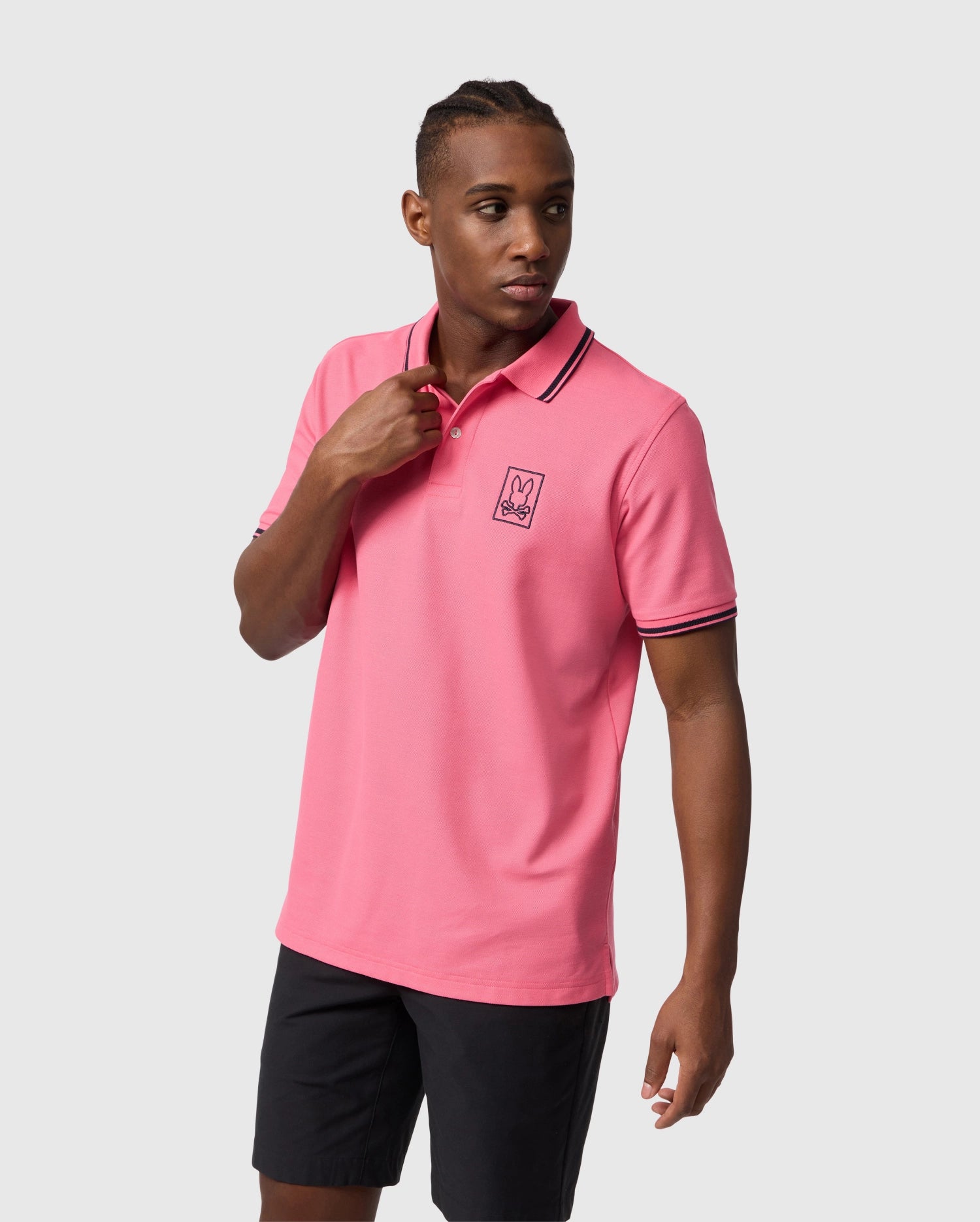 A young man in a Psycho Bunny MEN'S ARCADIA PIQUE POLO SHIRT - B6K406B200 with a crease-resistant collar, and a logo on the chest, paired with black shorts, looking to the side against a plain background.