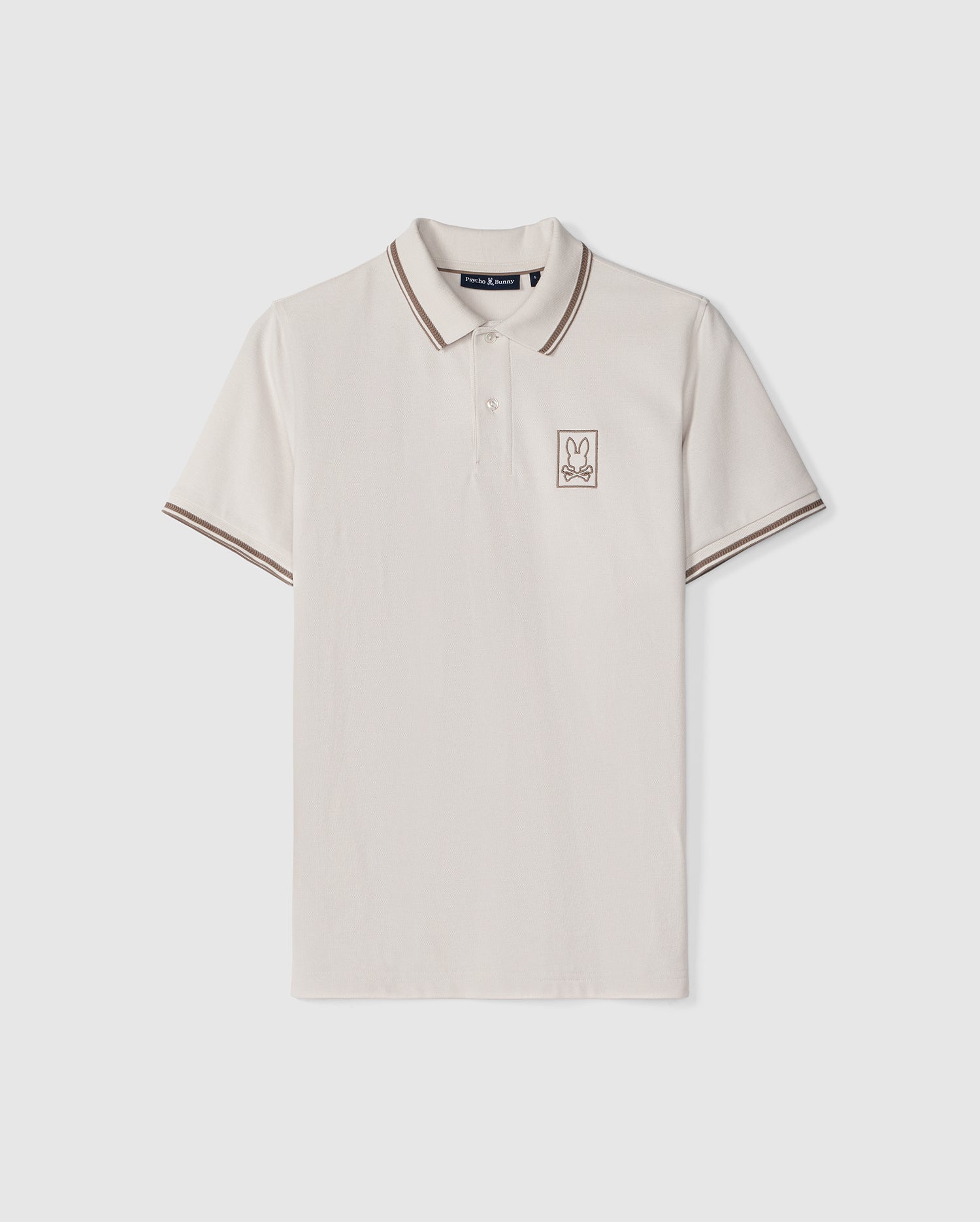 An Psycho Bunny polo shirt crafted from cream-colored Peruvian Pima cotton with brown trim on the crease-resistant collar and sleeves, featuring a small embroidered bunny logo on the left chest, displayed against a.