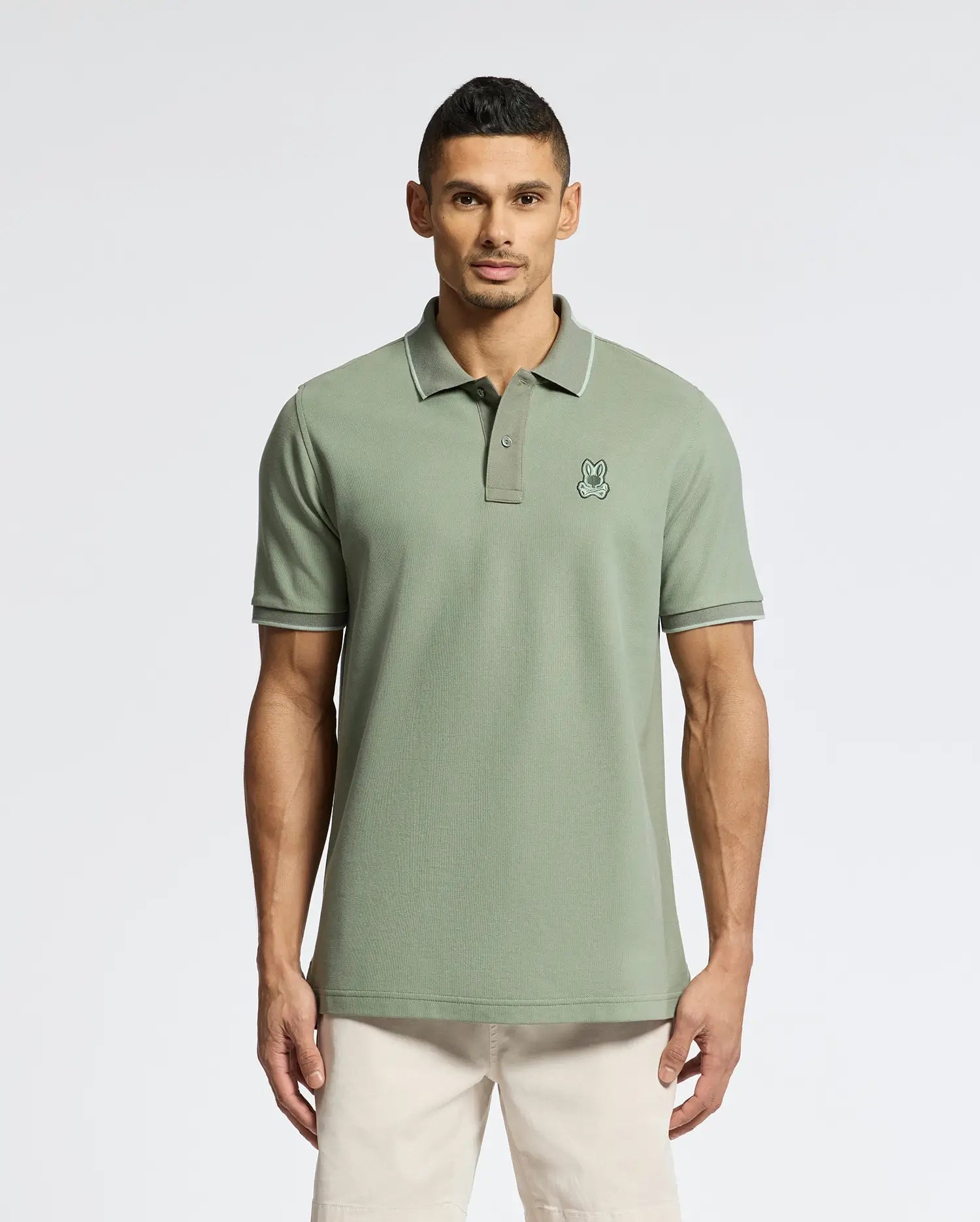 A man wears the Psycho Bunny MENS NEW CASTLE PIQUE POLO - B6K487F200 in green, a premium Pima cotton slim-fit shirt with a cartoon rabbit logo on the chest, standing against a plain white background to highlight its elegant contrast detailing.