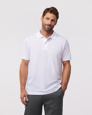 A man in a white pima cotton Psycho Bunny OUTLINE polo (B6K508ARPC) and grey pants stands against a plain background, looking at the camera with a slight smile. The polo features a small Psycho Bunny logo from the OUTLINE collection on the chest.