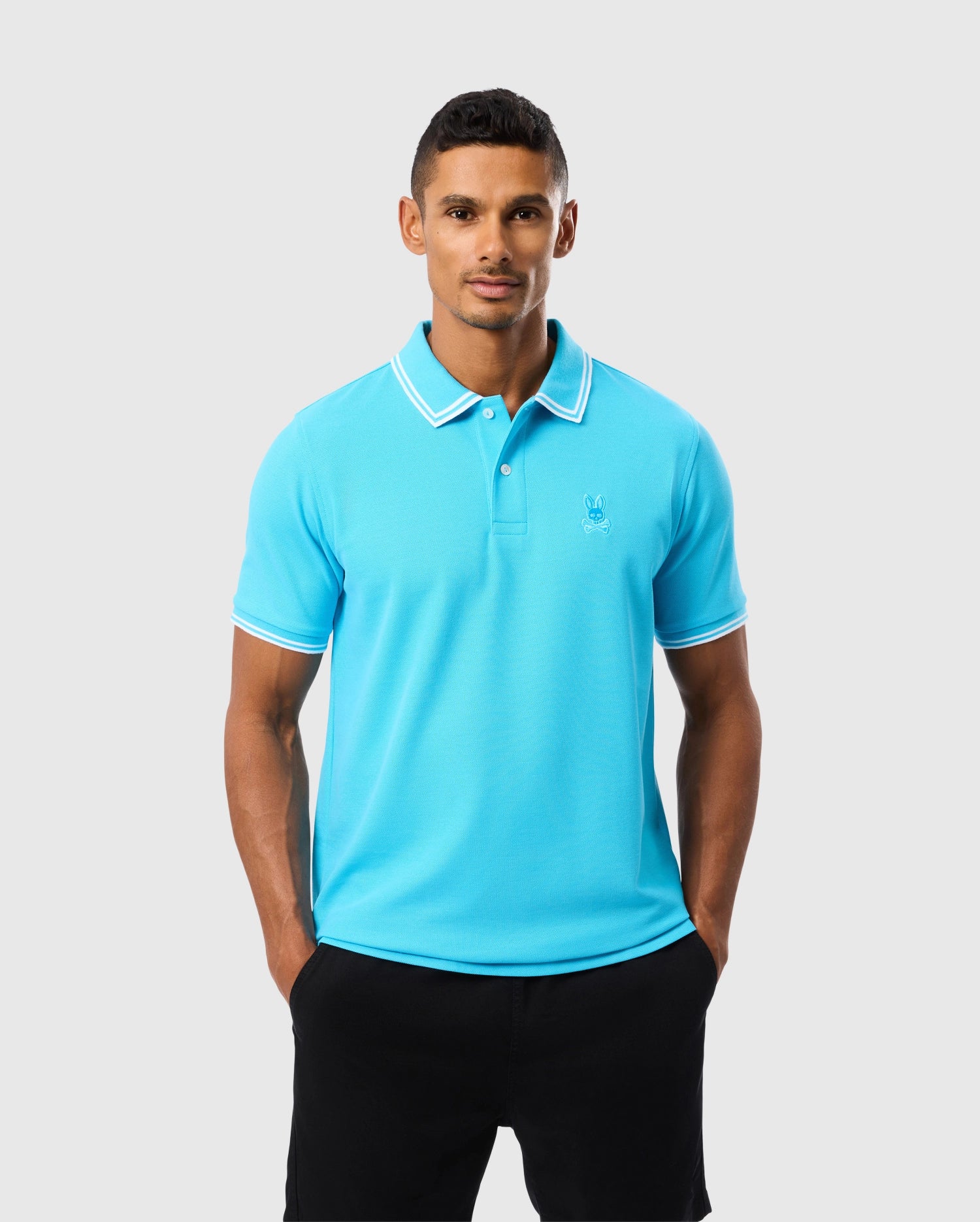 A man wearing a bright blue Psycho Bunny MENS BELTON PIQUE POLO - B6K575C200 with white trim on the collar and sleeves, and black shorts stands against a plain white background. He is looking directly at the camera with his hands in his pockets. The Pima cotton shirt features a small embroidered Bunny logo on the left chest.