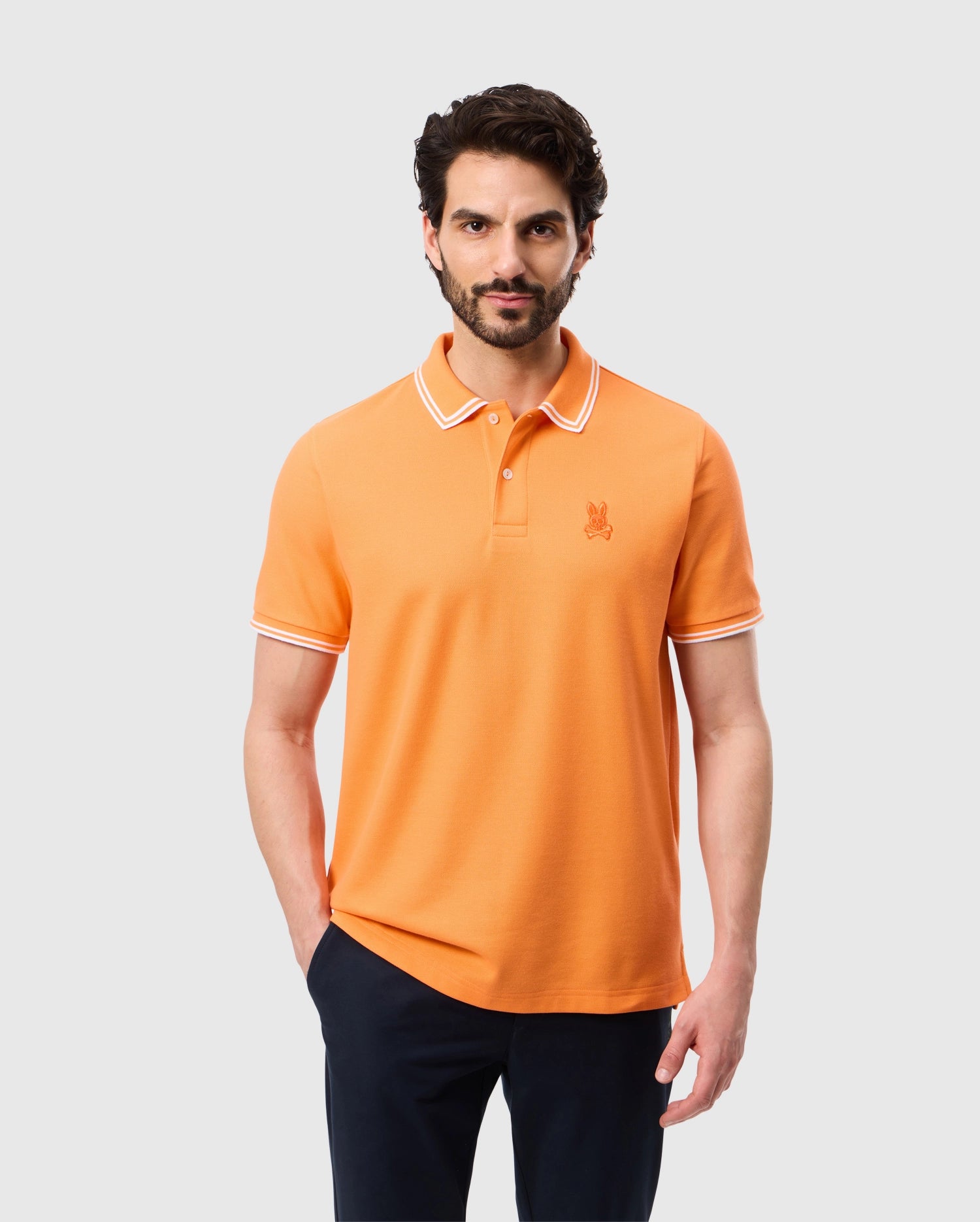 A man with dark hair and a beard is wearing an orange Psycho Bunny MENS BELTON PIQUE POLO - B6K575C200 with white trim on the collar and sleeves. The Pima cotton shirt features an embroidered Bunny logo on the chest. He stands against a plain white background with his hands in his pockets.