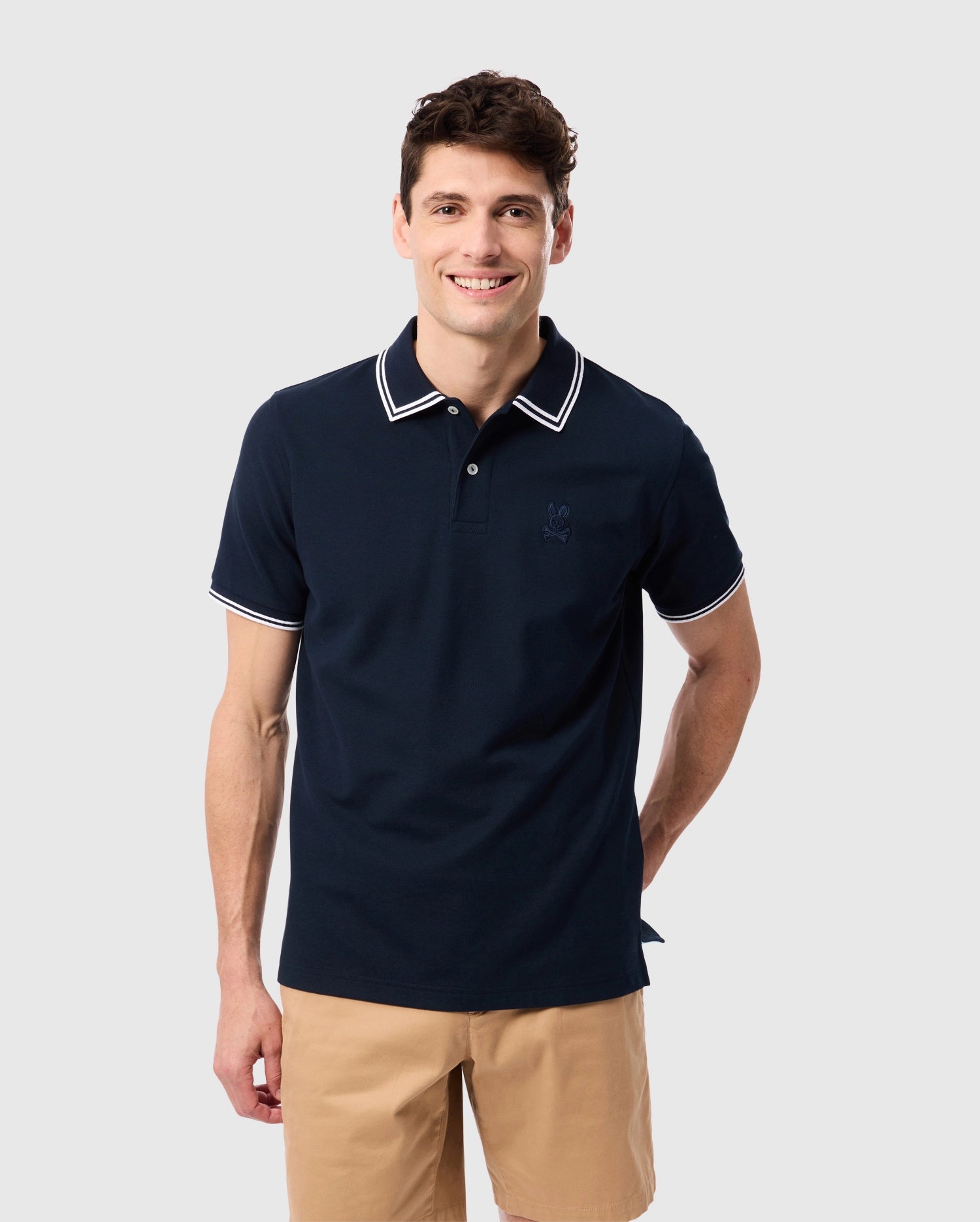 A man with short, dark hair is smiling and standing against a plain background. He is wearing a navy blue Psycho Bunny MENS BELTON PIQUE POLO - B6K575C200 with white, contrasting tipping on the collar and sleeves and light brown shorts. His left hand is in his pocket, while the right arm rests by his side.