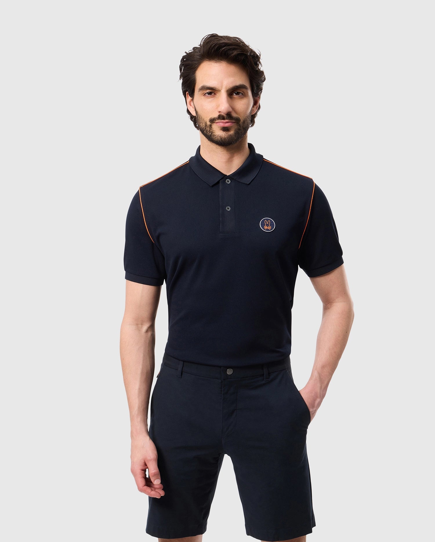 A man with dark hair and a beard is wearing a navy MENS WEBSTER SPORT POLO SHIRT - B6K580C200 by Psycho Bunny, featuring orange accents on the shoulders and a logo on the left chest. He is also wearing navy shorts. He stands against a plain light gray background with his hands by his sides.