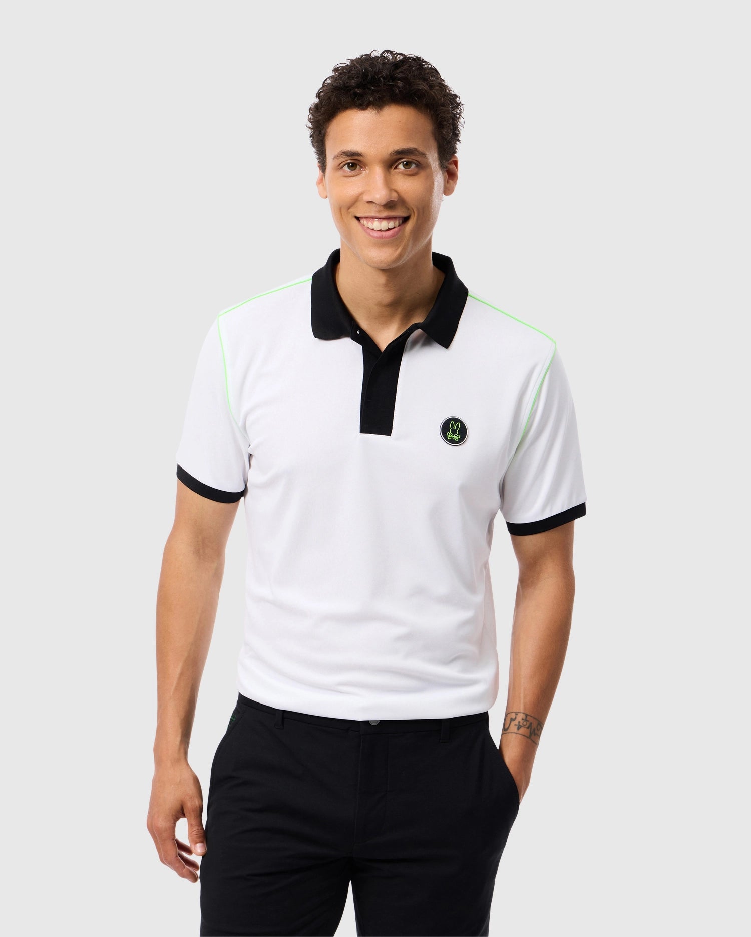 A person with short, curly hair stands smiling, wearing a Psycho Bunny MENS WEBSTER SPORT POLO SHIRT - B6K580C200. The shirt is white with a black collar, black sleeve cuffs, and features a small emblem on the chest. They also wear black pants. The background is a plain light gray color.