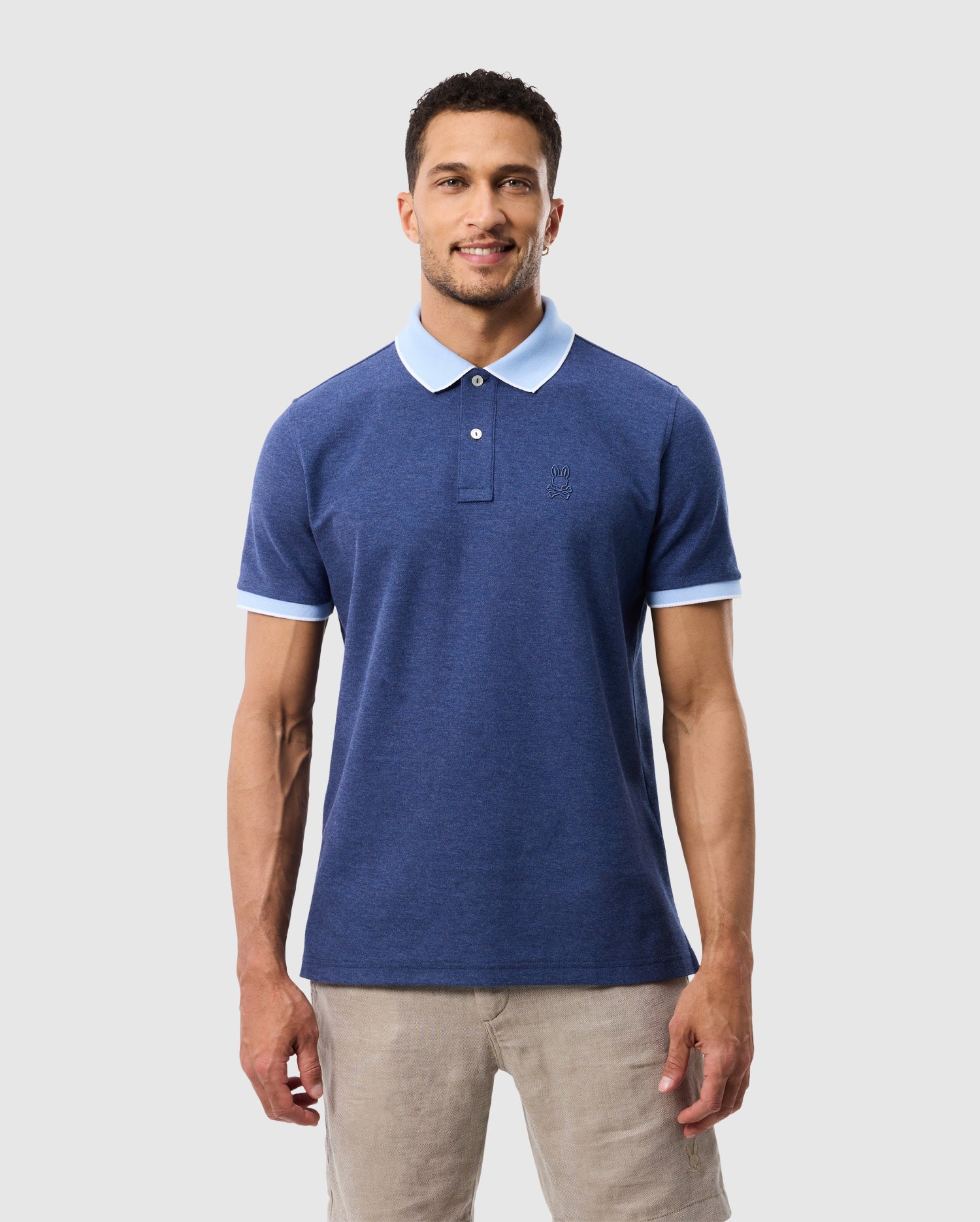A man with short hair is smiling and wearing a blue Psycho Bunny MENS WINDCREST PIQUE POLO SHIRT - B6K592C200 with light blue trim on the collar and sleeves. He is also donning beige pants, standing against a plain, light gray background.