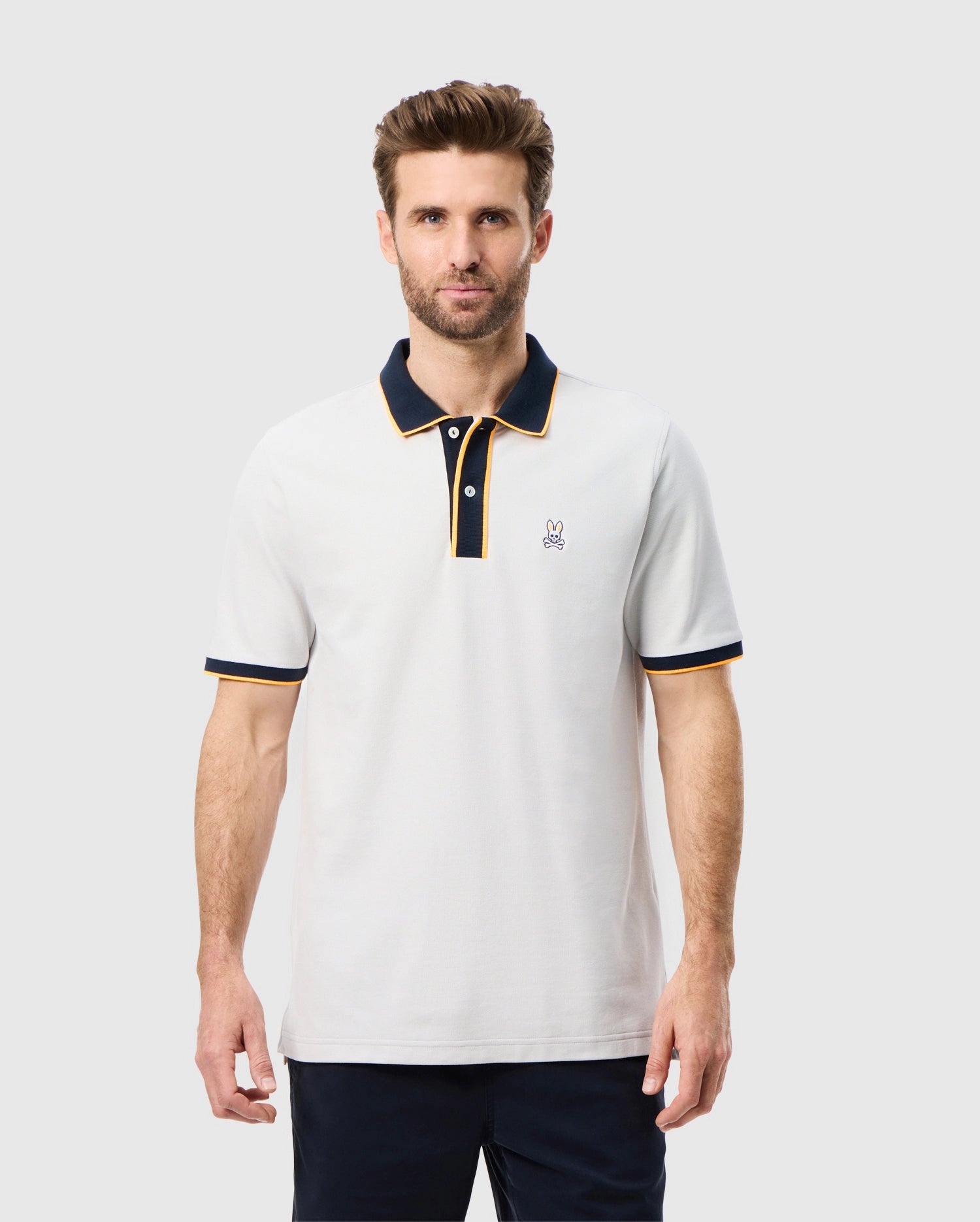 A man with brown hair and a beard is wearing a high quality cotton polo shirt in white, featuring black trim on the collar and sleeves. The MENS DALLAS PIQUE POLO SHIRT - B6K602C200 by Psycho Bunny has an embroidered Bunny logo on the left side of the chest. He is standing against a plain light gray background.