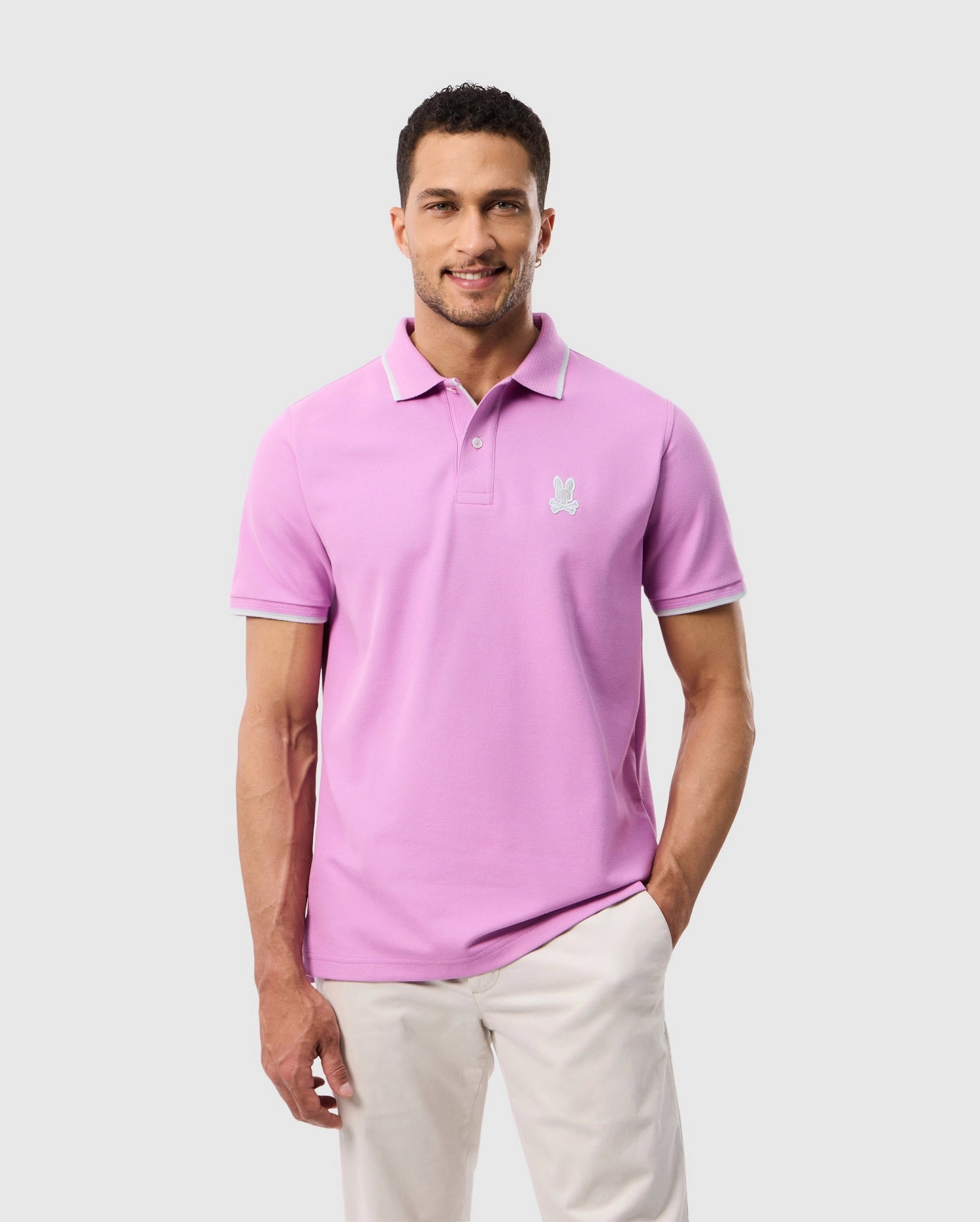 A man is wearing a light pink MENS HOUSTON PIQUE POLO SHIRT - B6K604C200 by Psycho Bunny with an embroidered bunny logo on the left side of his chest. He has short, dark hair and is posing against a plain, light grey background with one hand in his pocket. He is also wearing light-colored pants.