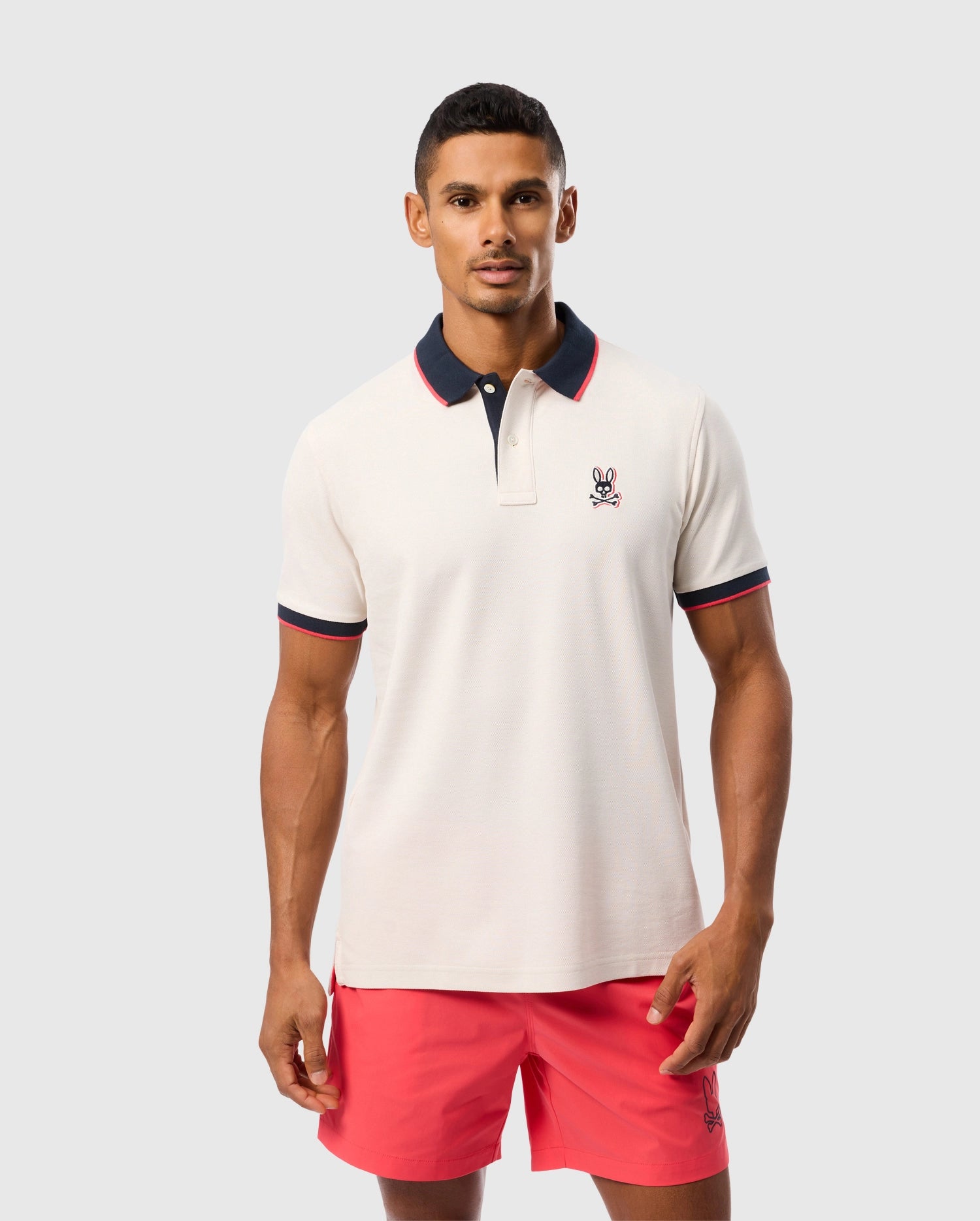 A man stands against a plain background wearing a Psycho Bunny MENS KAYDEN PIQUE POLO - B6K675C200 with a navy collar and red trim, featuring a small rabbit logo on the left chest. He is also sporting pink shorts with a small rabbit logo at the hem.
