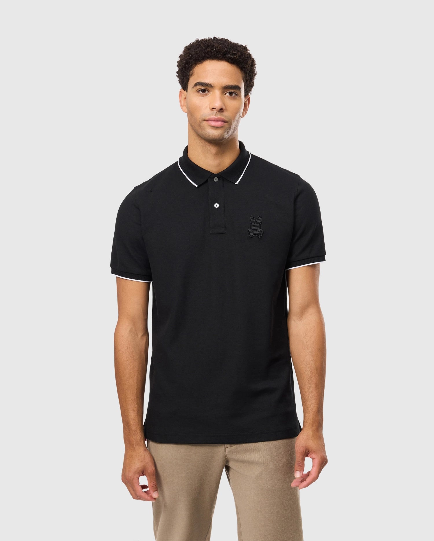 A person wearing the Psycho Bunny MENS KINGSTON PIQUE POLO SHIRT - B6K764D200 in black with white trim stands against a plain light gray background. This polo, crafted from soft Pima cotton, showcases a subtle embroidered Bunny patch on the chest. Their look is completed with stylish tan pants.