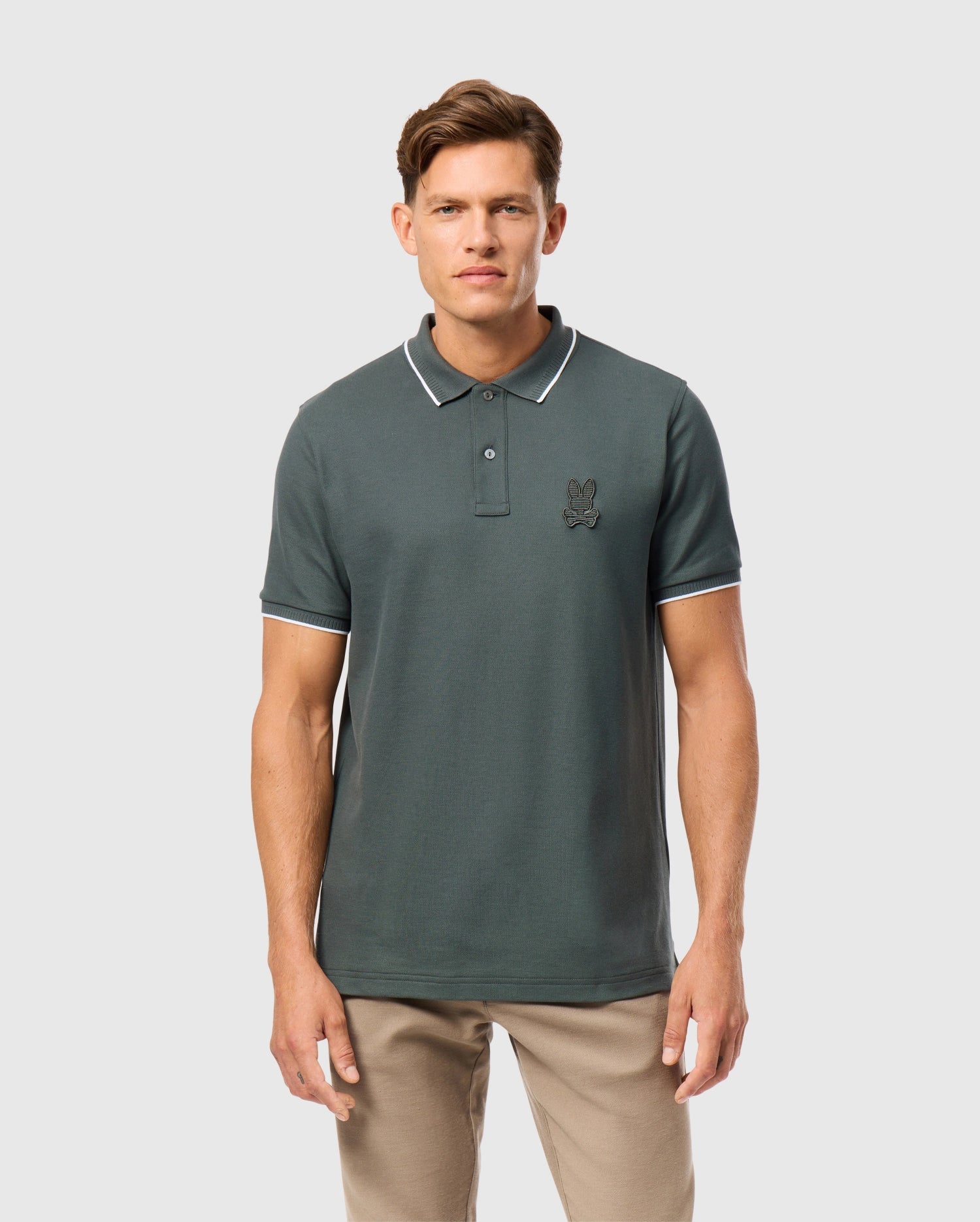 A man with short brown hair is wearing a dark green MENS KINGSTON PIQUE POLO SHIRT - B6K764D200 by Psycho Bunny, made of Pima cotton, featuring light-colored trim and an embroidered Bunny patch on the left chest. He is also dressed in beige pants and standing against a light grey background.