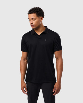 A person with short, curly hair is wearing a black MENS BROADWAY PIMA INTERLOCK JOHNNY COLLAR POLO by Psycho Bunny. This polo, made from durable Mercerized Pima cotton, features a subtle chest logo. Paired with black pants, they're standing in front of a plain light gray background, gazing off to the side.