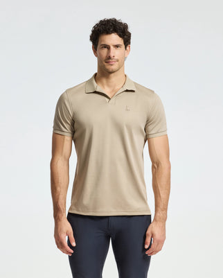 A man with curly hair is wearing the Psycho Bunny MENS BROADWAY PIMA INTERLOCK JOHNNY COLLAR POLO - B6K891D200 in light brown, featuring a small emblem, paired with dark pants, standing against a plain white background.