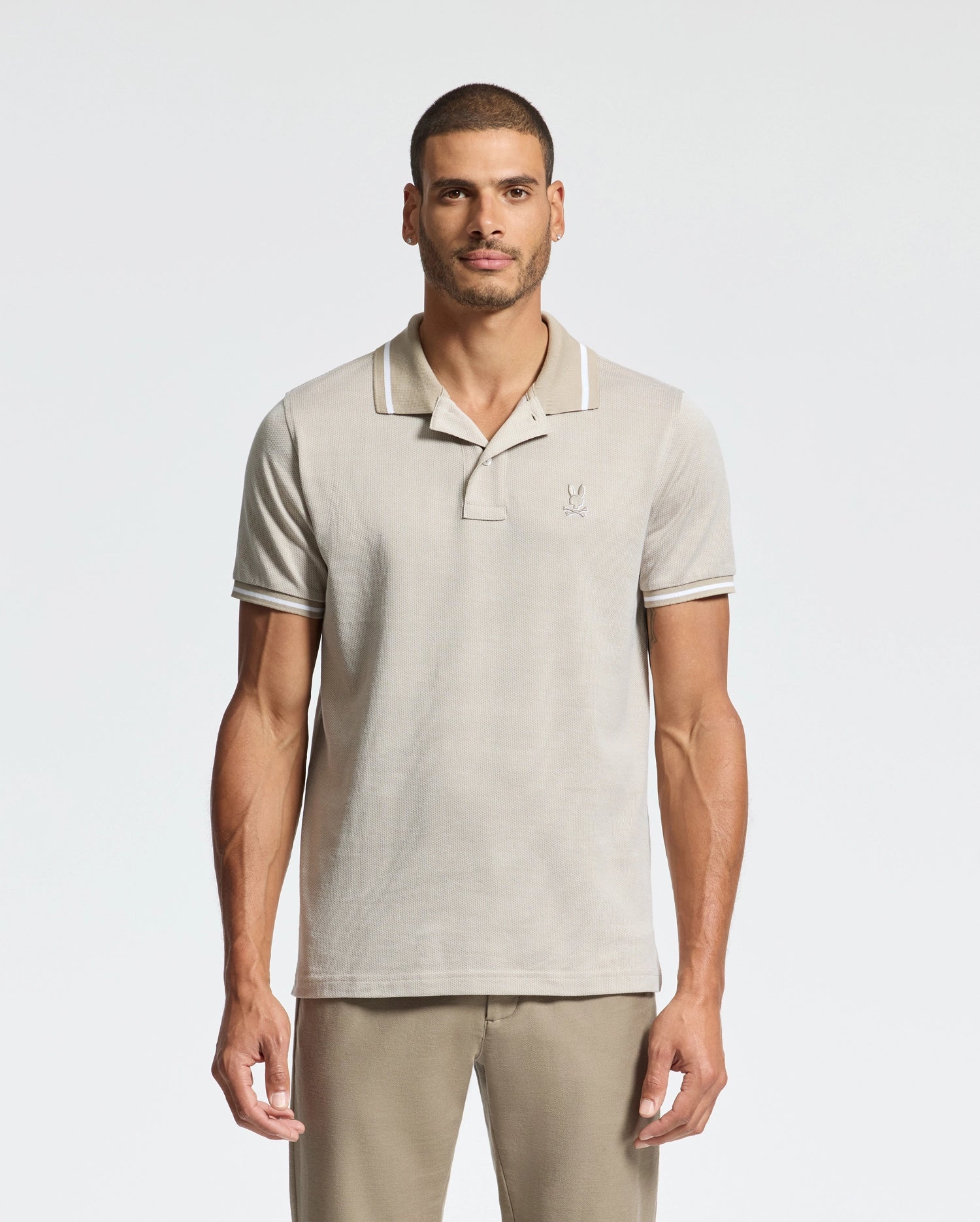 A man wearing a light khaki Psycho Bunny Men's Warsaw Jacquard Pique Polo, crafted from Peruvian Pima cotton, stands against a plain white background. The shirt features dark trim on the collar and sleeves, along with a small embroidered logo. He has short hair and a trimmed beard, with his hands by his sides.