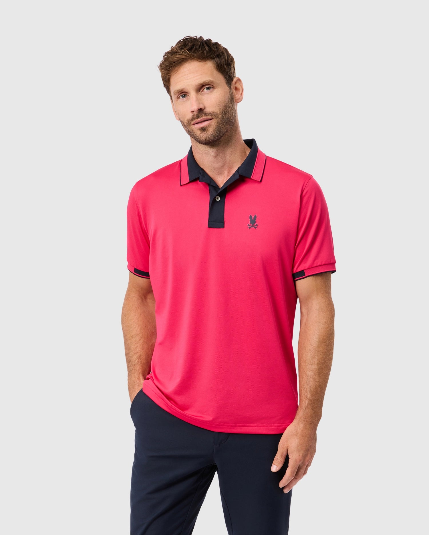 A man with brown hair and a beard is wearing the Psycho Bunny MENS BELLEVUE SPORT POLO SHIRT - B6K905D200 in bright pink, featuring a small emblem on the chest, paired with dark navy trousers. He stands against a plain light gray background, looking at the camera with a neutral expression.