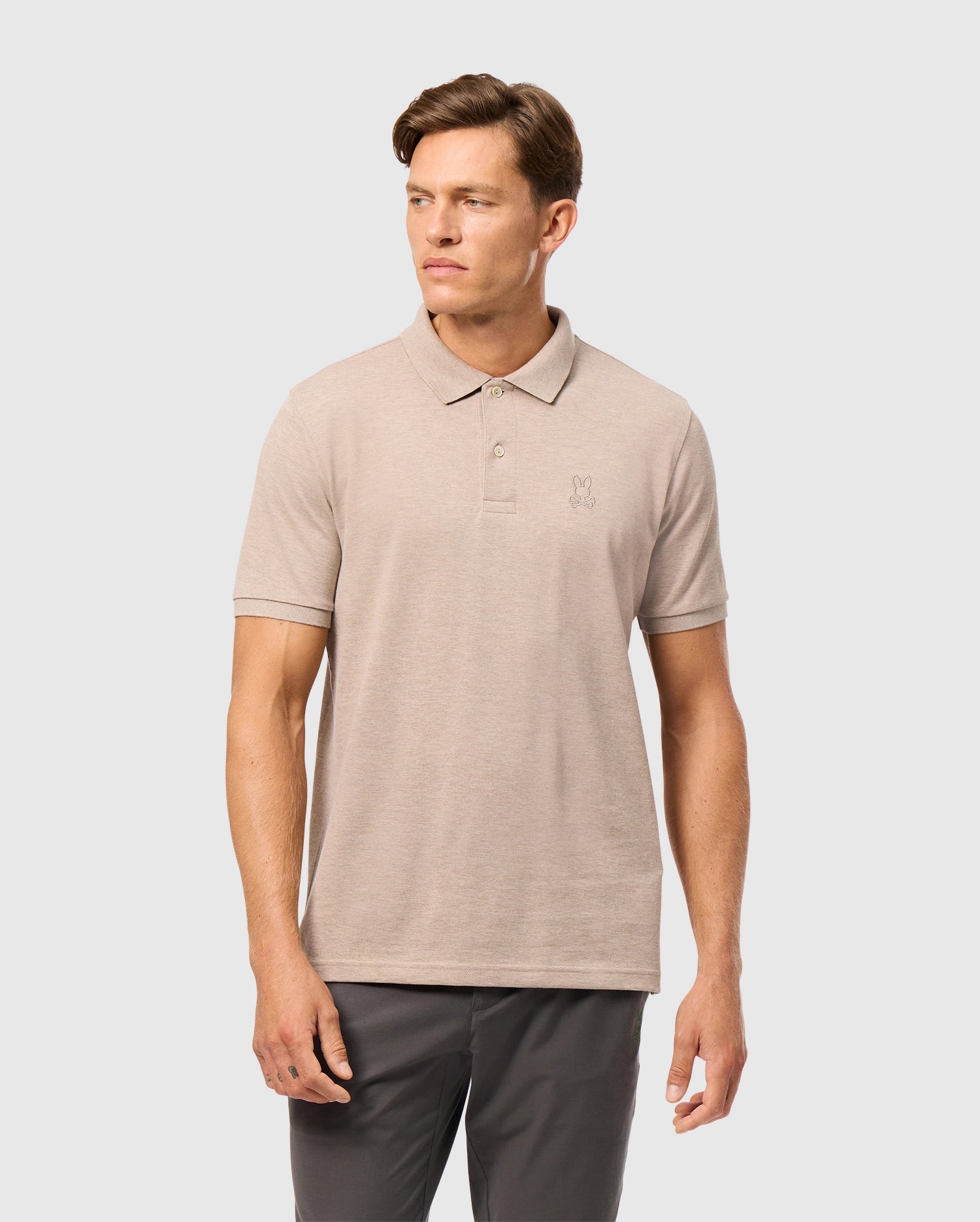 A man in a Psycho Bunny MENS JAMES PREMIUM PIQUE POLO SHIRT - B6K945D200, made of lightweight and durable Pima cotton featuring a small embroidered logo on the chest, stands against a plain gray background, looking off to the side.