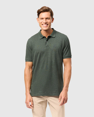 A person in a Psycho Bunny MENS JAMES PREMIUM PIQUE POLO SHIRT (B6K945D200) and beige pants smiles while standing against a plain gray background.