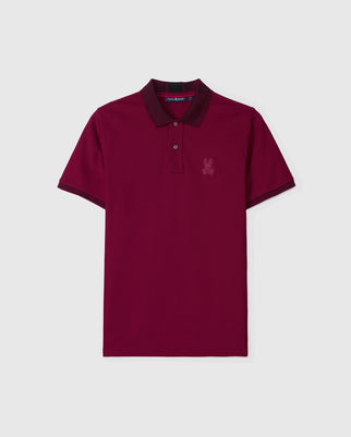 The Psycho Bunny Men's Malton Pique Polo Shirt - B6K950D200 is a classic fit polo made from premium Pima cotton, ensuring both comfort and durability. This stylish short sleeve shirt is maroon with a dark collar and sleeve trim, featuring a subtle embroidered logo on the chest, and is displayed against a light gray background.