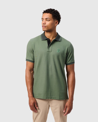 A person wearing a Psycho Bunny's MENS MALTON PIQUE POLO SHIRT - B6K950D200, characterized by its luxurious pima cotton and subtle pique pattern in green with contrasting dark collar and sleeve trim, stands against a plain white background. The polo features a small embroidered logo on the left side. The individual is looking to the side and is paired with beige pants.