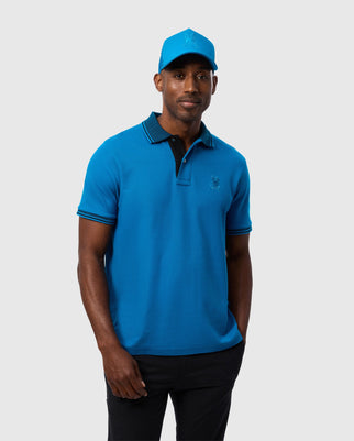 A man is standing against a plain background, wearing a vibrant bright blue Psycho Bunny MENS MALTON PIQUE POLO SHIRT - B6K950D200 made from Pima cotton, featuring a small embroidered logo on the chest. He is also wearing a matching blue cap and gazes at the camera with a relaxed expression, his left hand in his pocket.