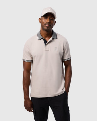 A man wearing a Psycho Bunny MENS MALTON PIQUE POLO SHIRT - B6K950D200 in light gray, embellished with black collar and sleeve trim, along with a matching light gray baseball cap, stands against a plain gray background. The shirt features an embroidered emblem on the chest and has a subtle diamond pique pattern; he casually has one hand in his pocket.