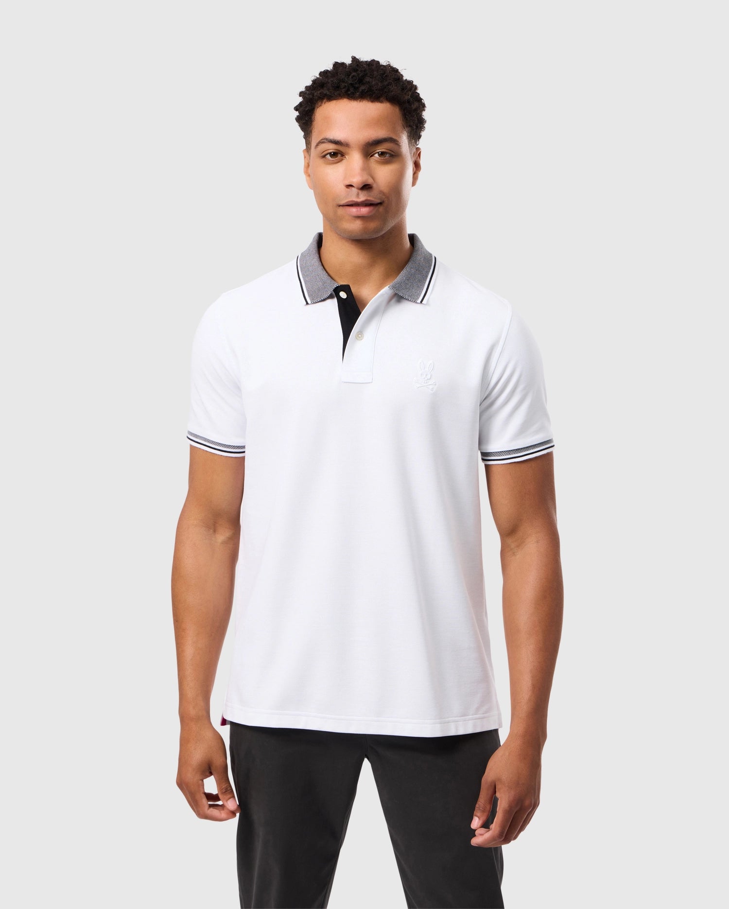 Standing against a simple gray background, a man showcases the Psycho Bunny Men's Malton Pique Polo Shirt (B6K950D200). This white polo features a black collar and sleeve trim, crafted from sleek Pima cotton with elegant mother-of-pearl buttons, exemplifying the distinctive Psycho Bunny style.