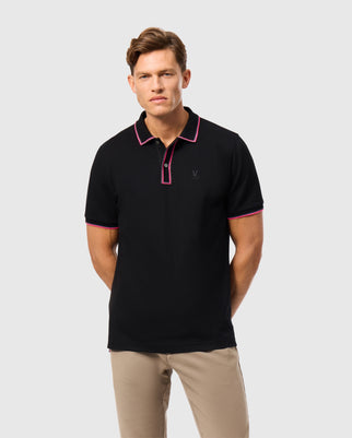 A man wearing the Psycho Bunny Men's Kent Pique Polo Shirt, featuring a sleek black design with pink trim and a small embroidered logo on the left chest, stands against a plain background. His hands are in his pockets, he has short brown hair, and he pairs the polo with beige trousers.