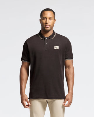 A man is standing against a plain backdrop, wearing the Psycho Bunny MENS MONROE PIQUE POLO - B6K964D200. The polo is black Pima cotton with beige trim and mother-of-pearl buttons, featuring a small embroidered patch on the chest. He has short hair and is paired with beige pants.